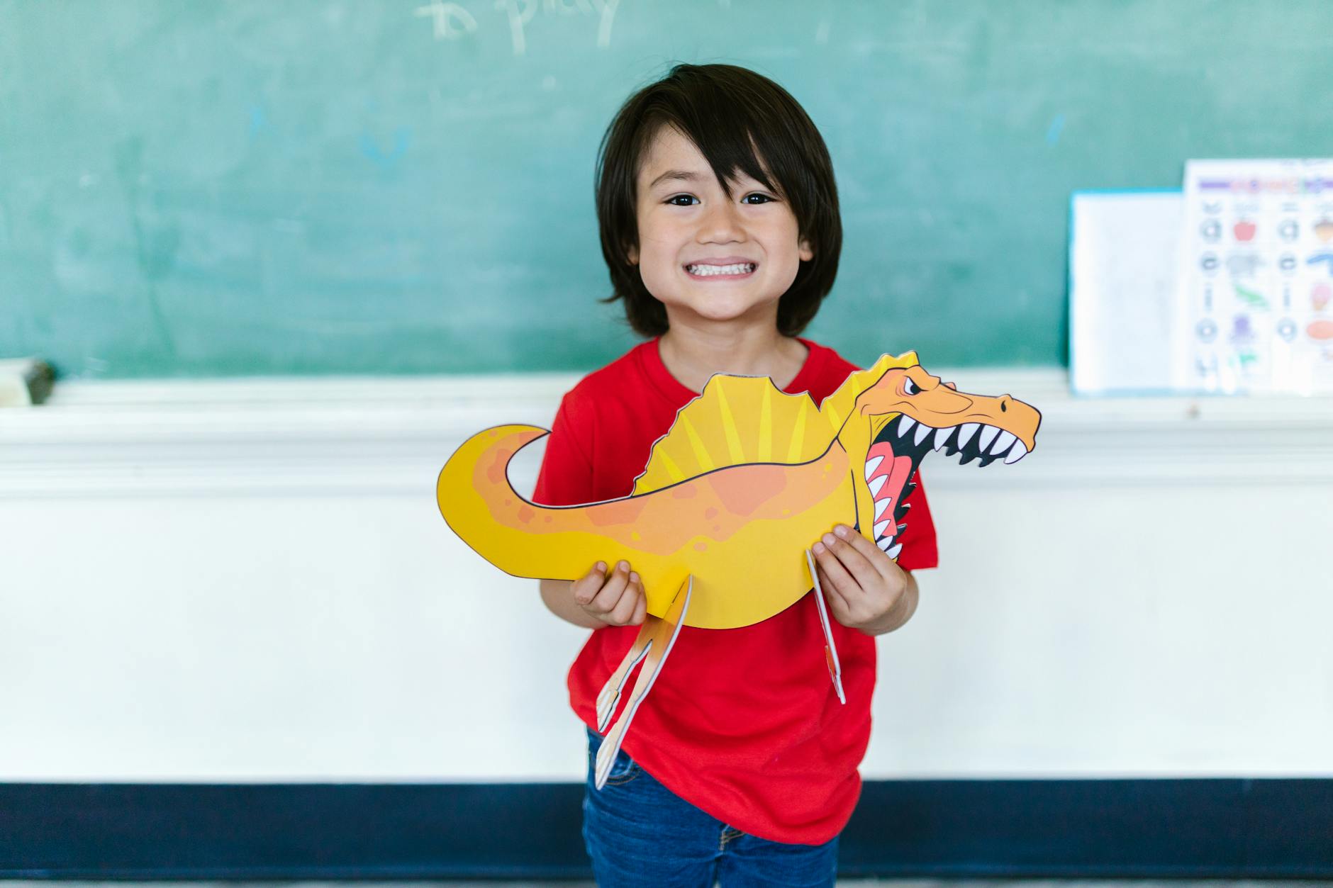 A cheerful child holds a colorful dinosaur cutout in a classroom setting. - school closure activities