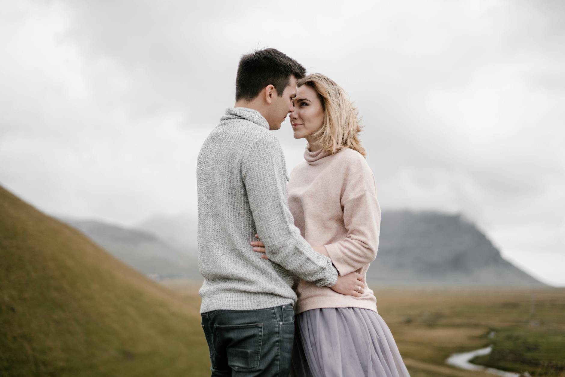 Side view glad young couple in warm sweaters embracing and looking at each other while spending time together on grassy hillside valley against majestic mountain and river on cold cloudy day - secure attachment style