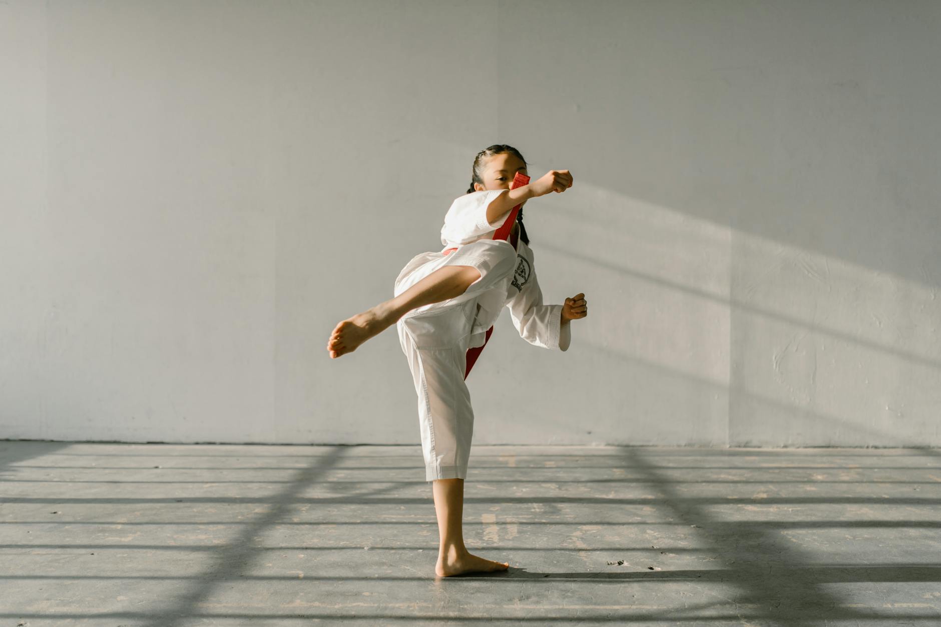 Asian girl in martial arts uniform practicing a high kick indoors during daytime. - self-compassion practices