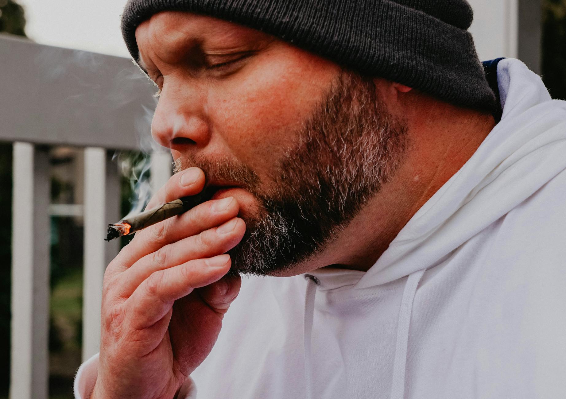 A bearded man in a hoodie and beanie smoking a hand-rolled cigarette outdoors in daylight. - setting boundaries adult children