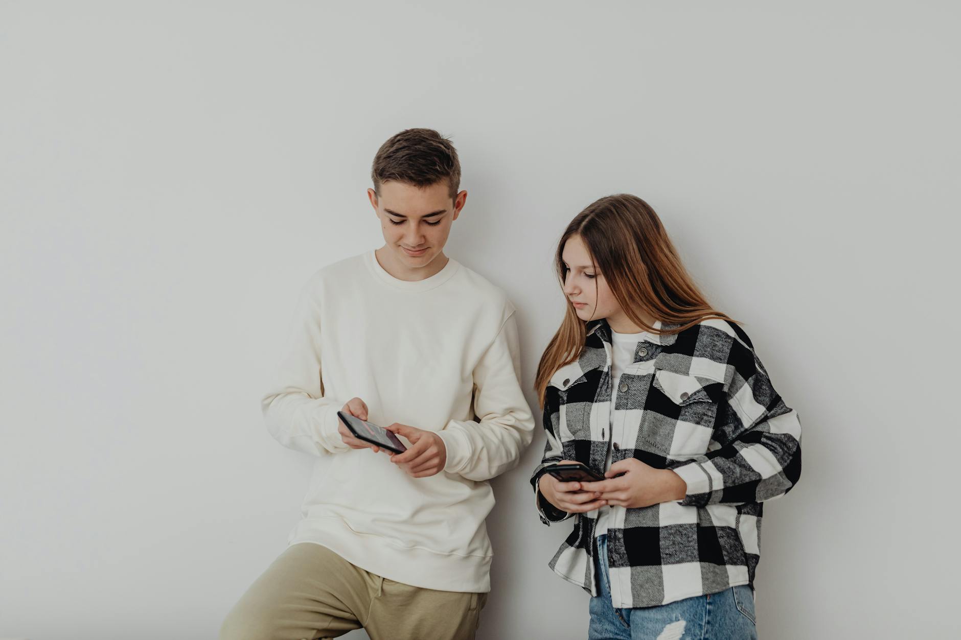 Teenage boy and girl using smartphones while leaning against a white wall indoors. - social media effects on teens