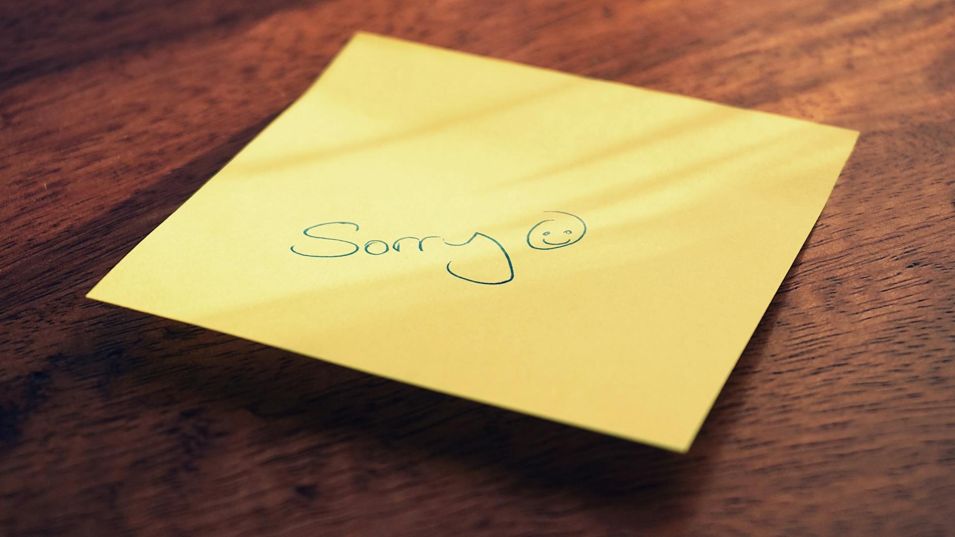 A yellow sticky note with a handwritten apology message and smiley face on a wooden surface. - how to spell apologize
