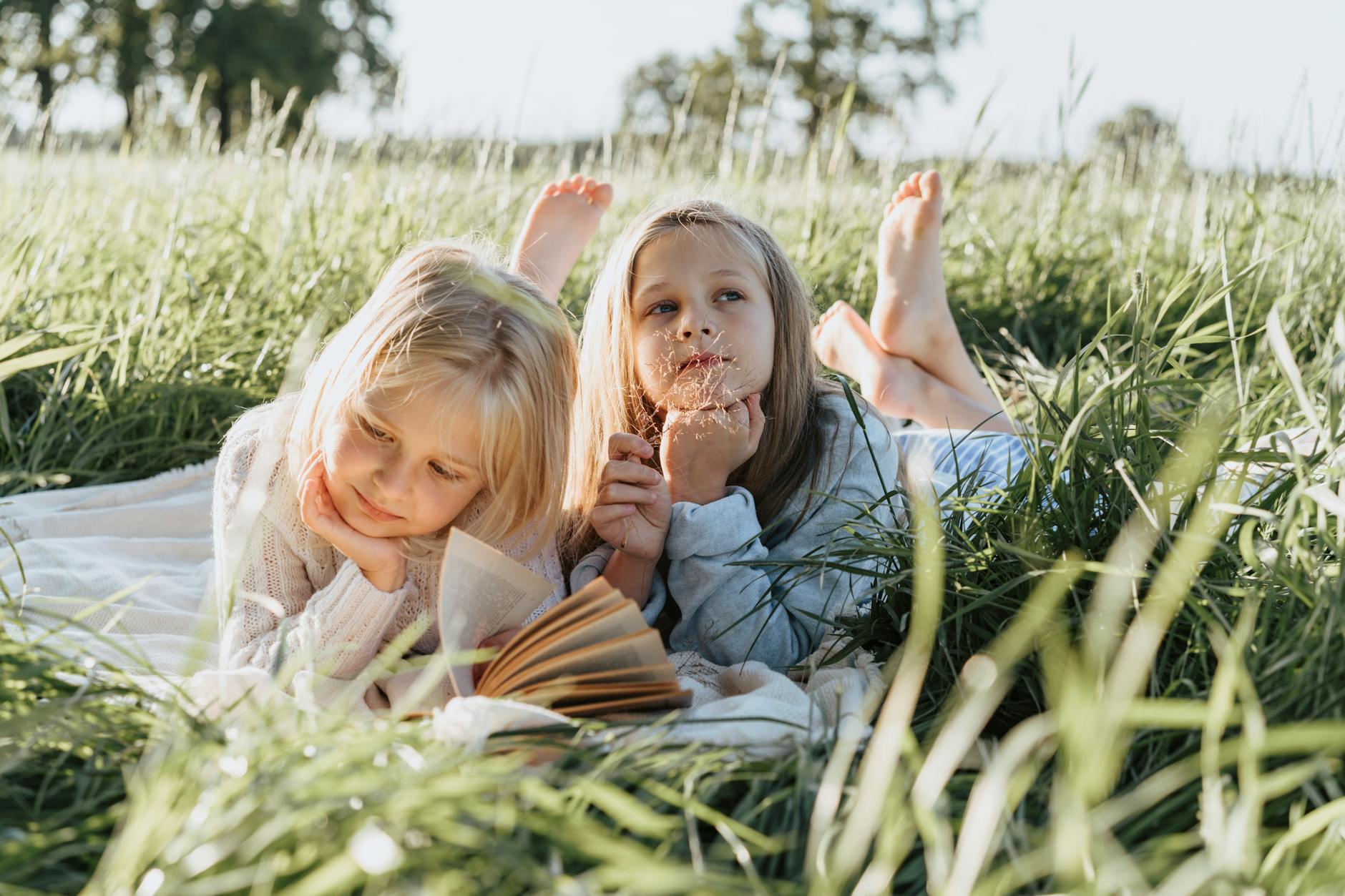 Two young girls lying on a picnic blanket in a sunny field, reading and daydreaming. - spring break kids