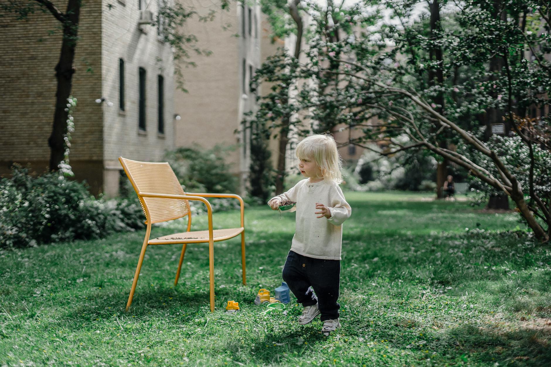 Full body of adorable little boy in stylish outfit walking on grassy lawn near yellow chair in backyard on sunny day - spring child energy