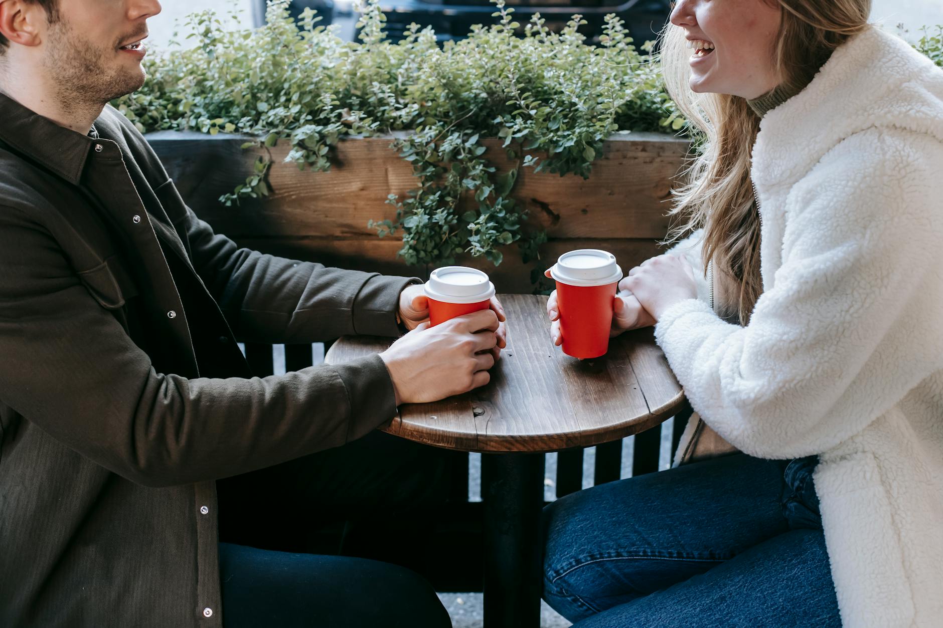 Crop faceless couple sitting at wooden table with paper cups of coffee and looking at each other while having date - spring dating refresh