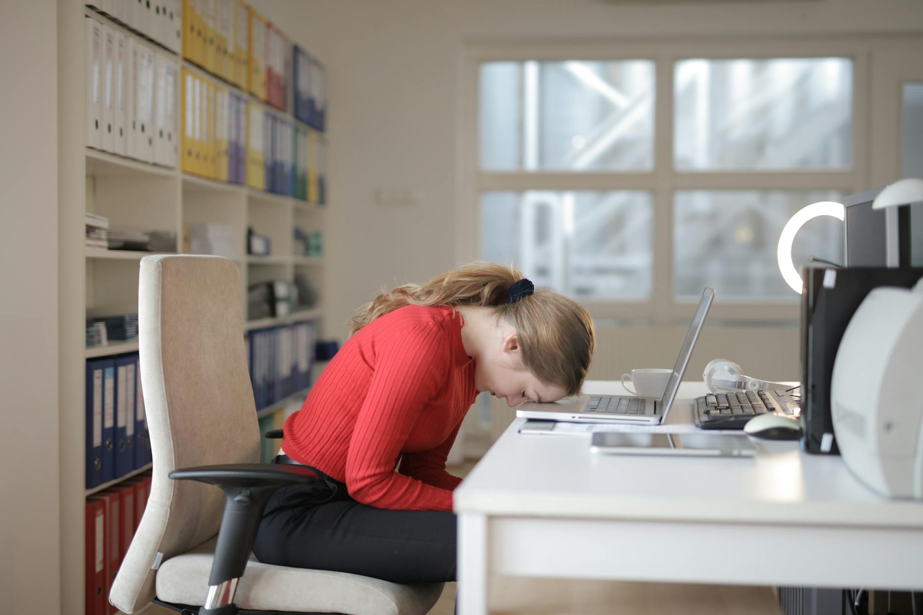 Tired woman in red sweater naps on office desk beside laptop, overwhelmed by remote work. - spring fatigue causes