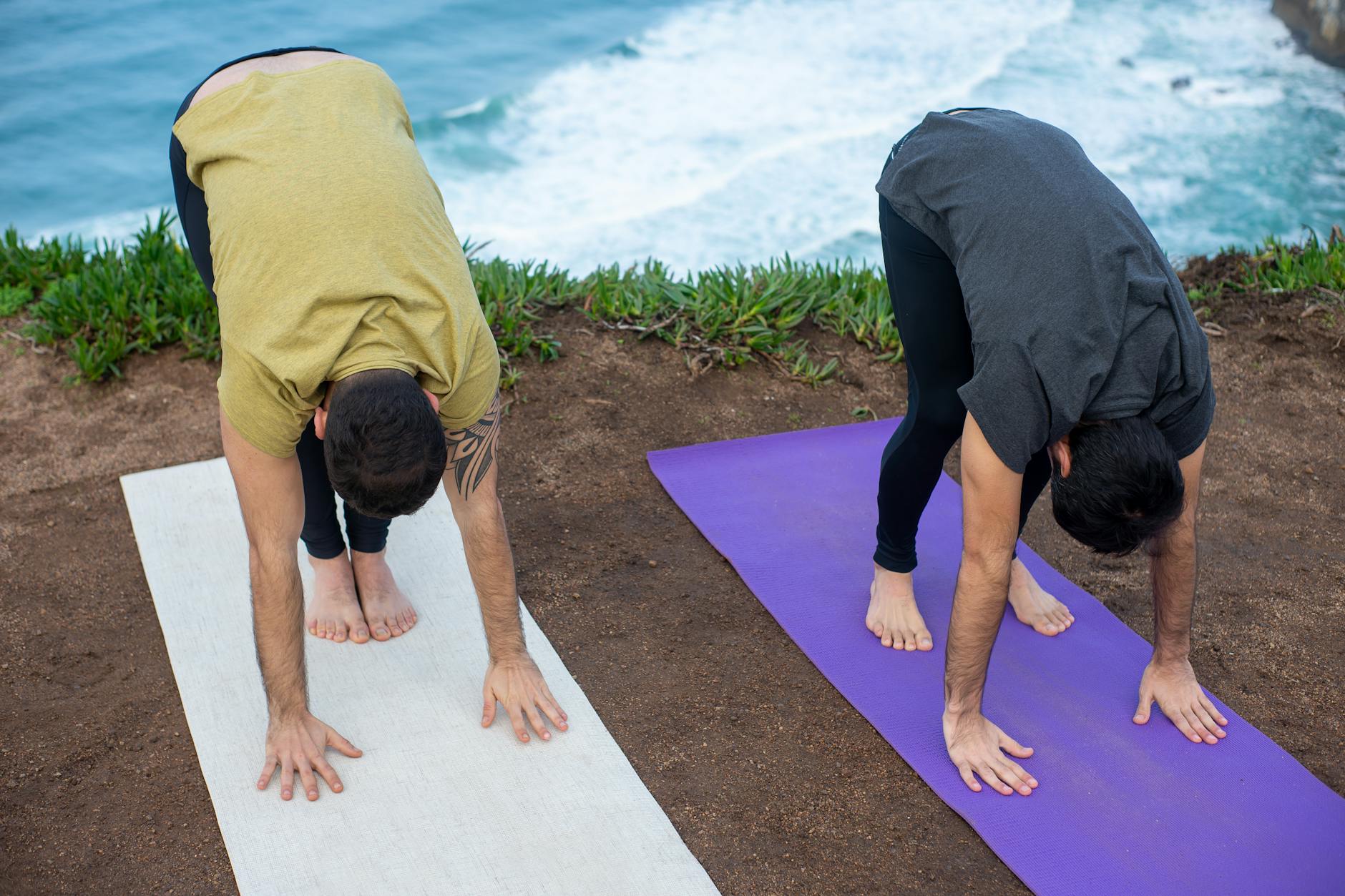 Two men practicing yoga outdoors by the sea in Portugal, performing forward bends on yoga mats. - spring forward mental health