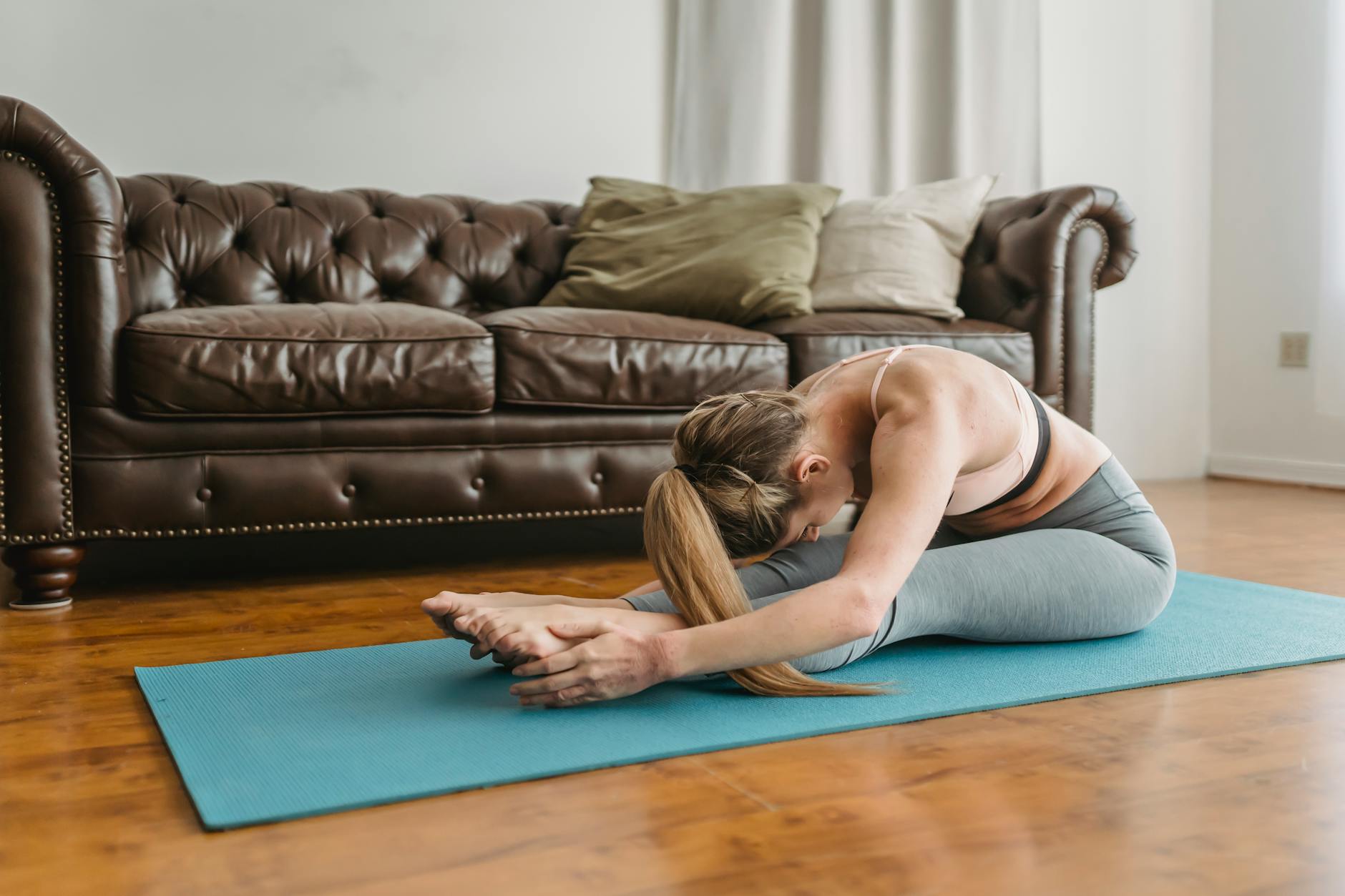 Side view of slim young lady with long blond hair in sportswear recreating in Seated Forward Bend asana after yoga session in living room - spring forward mental health