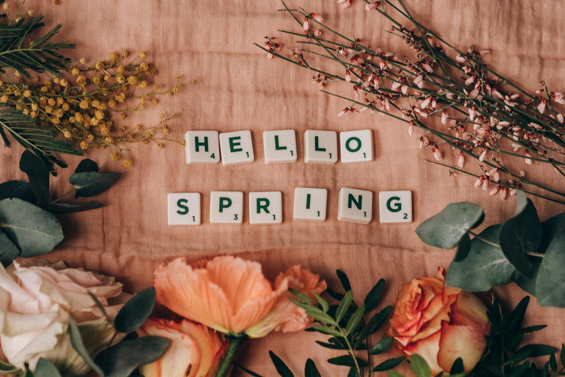 A spring-themed floral arrangement with Scrabble tiles spelling 'HELLO SPRING' on a pink surface. - spring mood boost