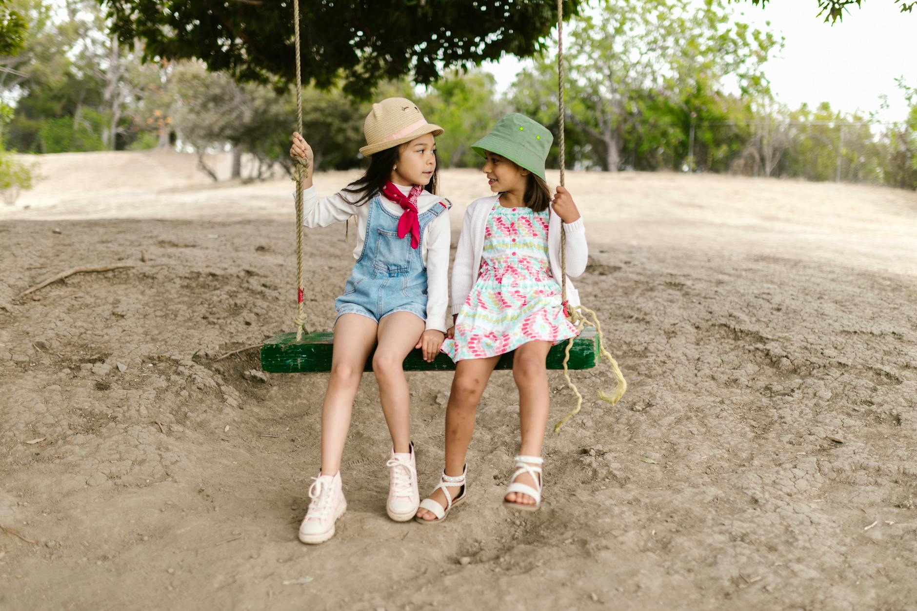 Two young girls in bucket hats share a joyful swing ride in a lush outdoor setting. - spring mood swings