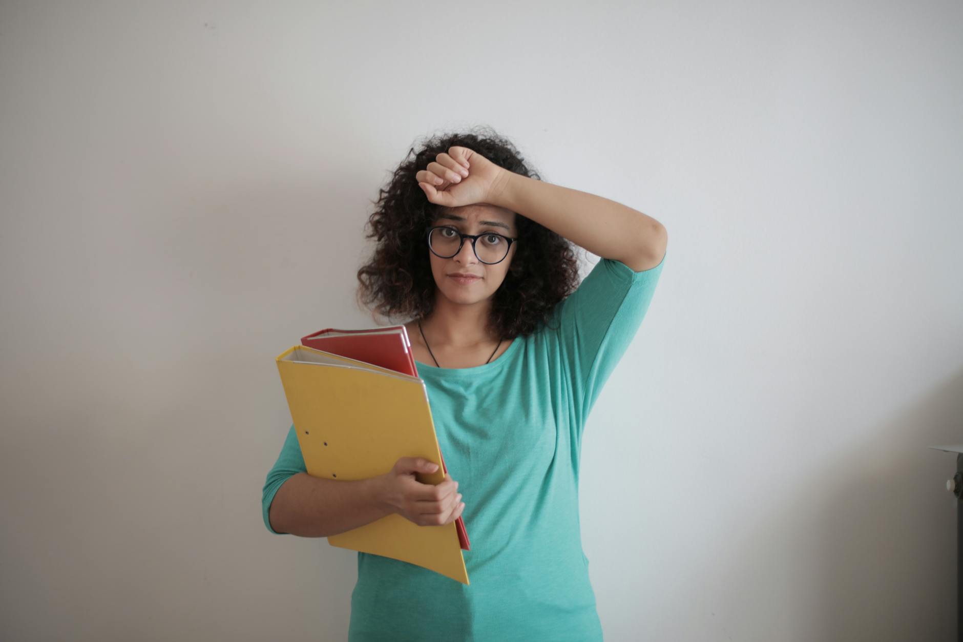 Unhappy overworked female employee in glasses and casual clothes with folders of documents looking at camera with sadness and touching head while standing against white wall in contemporary office - spring overwhelm therapy
