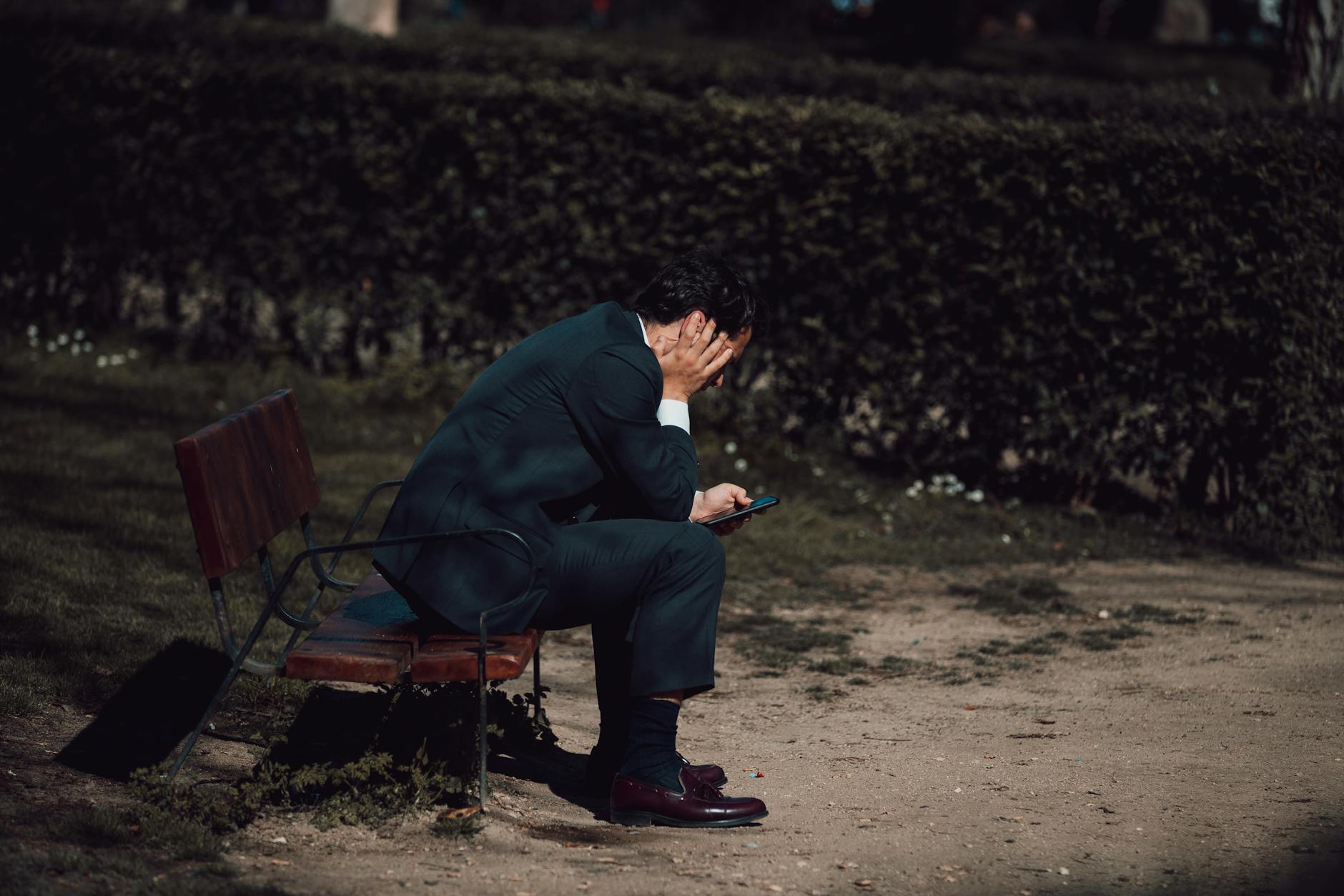 A worried businessman sits on a park bench in Madrid, holding a smartphone. Captured outdoors in natural light. - spring overwhelm therapy