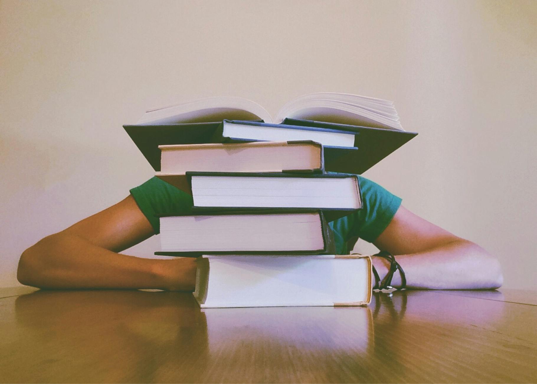 A student hidden behind a stack of books, symbolizing academic stress and study challenges. - spring overwhelm therapy