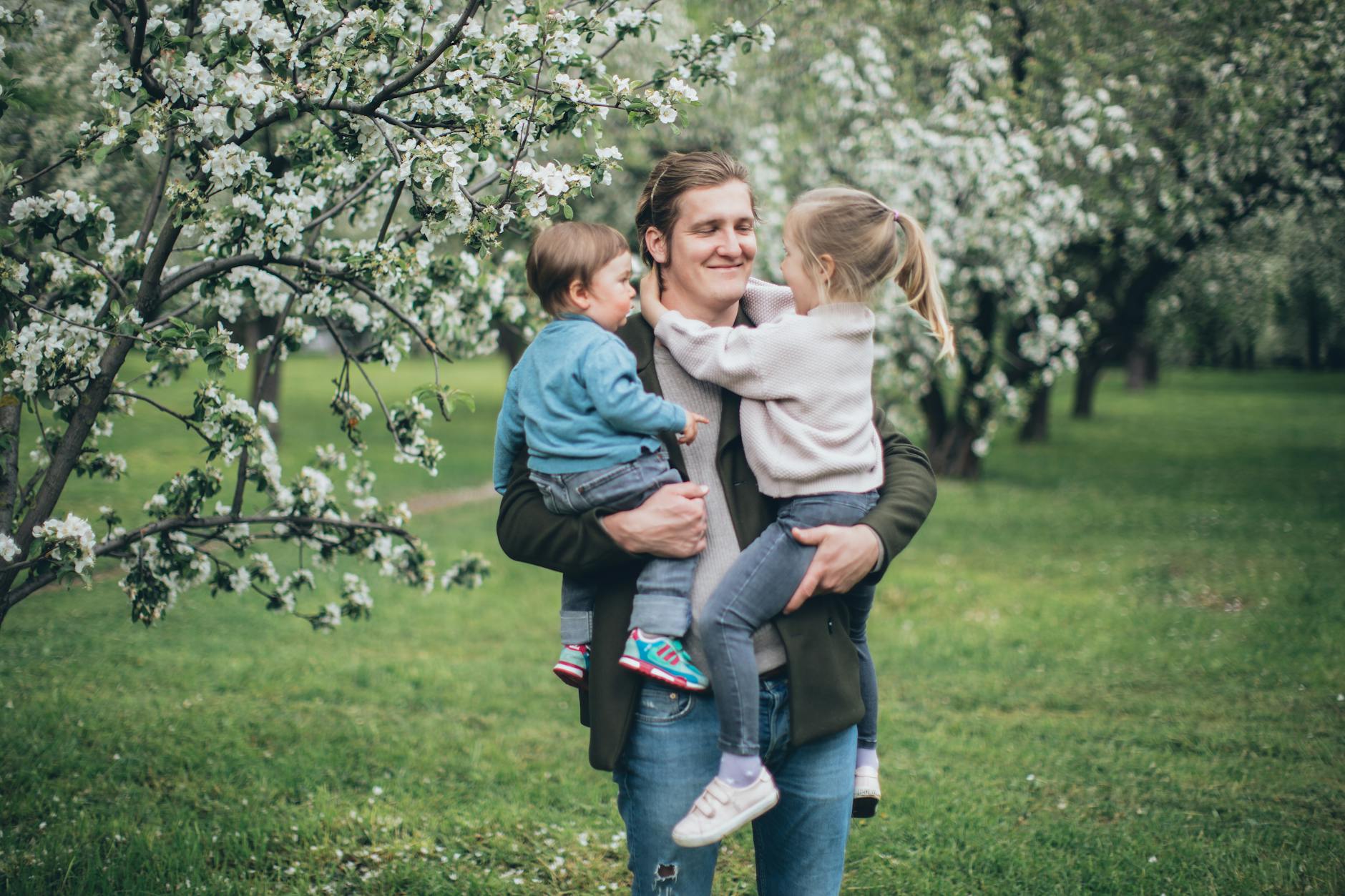 A loving father holds his children among blooming spring trees. - spring parenting tips