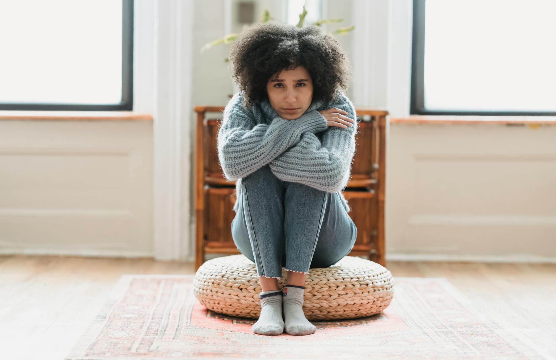 Full body of sad African American female looking at camera while sitting on rattan mat in light room at home - spring sad symptoms