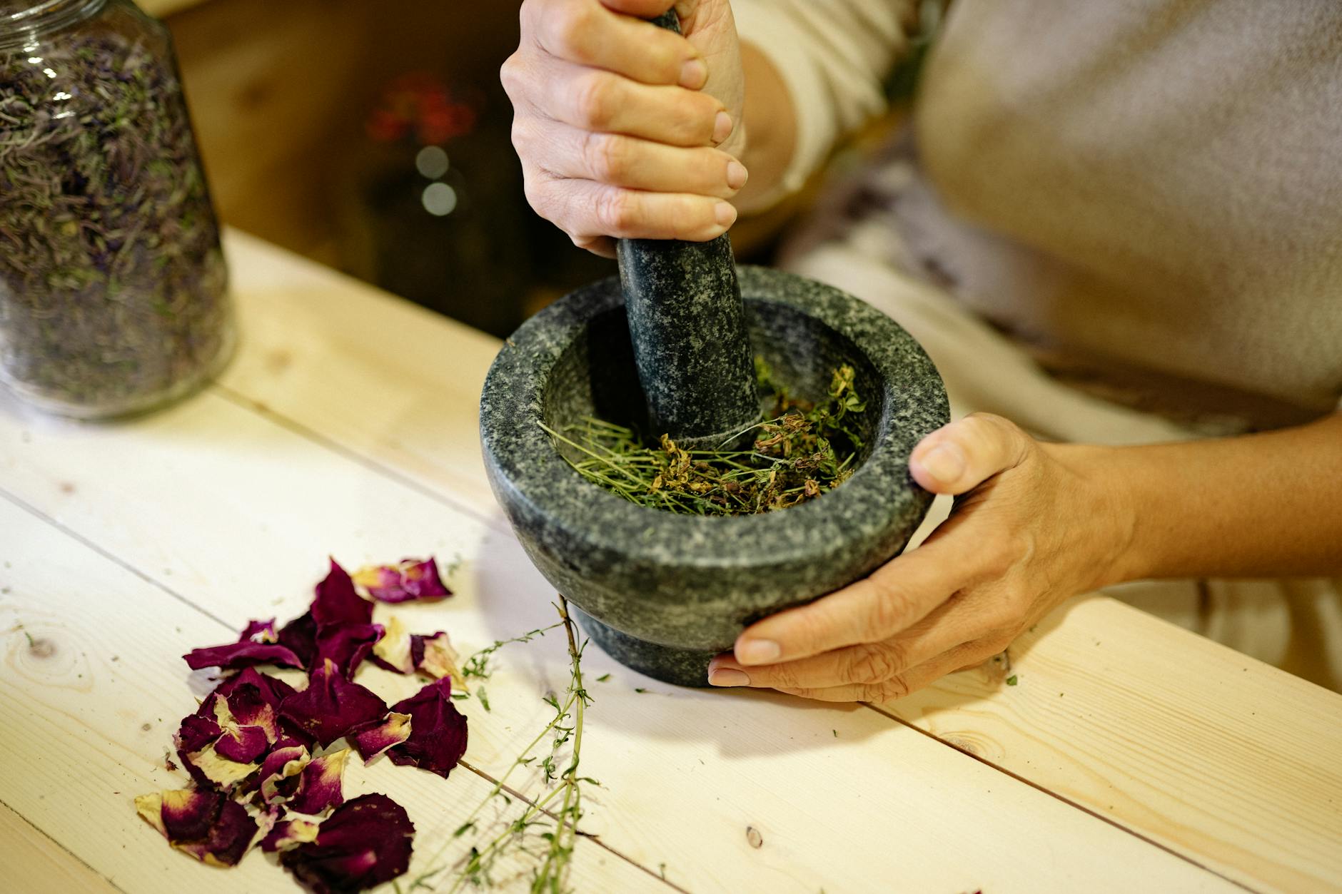 A person grinding herbal ingredients in a stone mortar and pestle with dried petals nearby. - spring slump remedies
