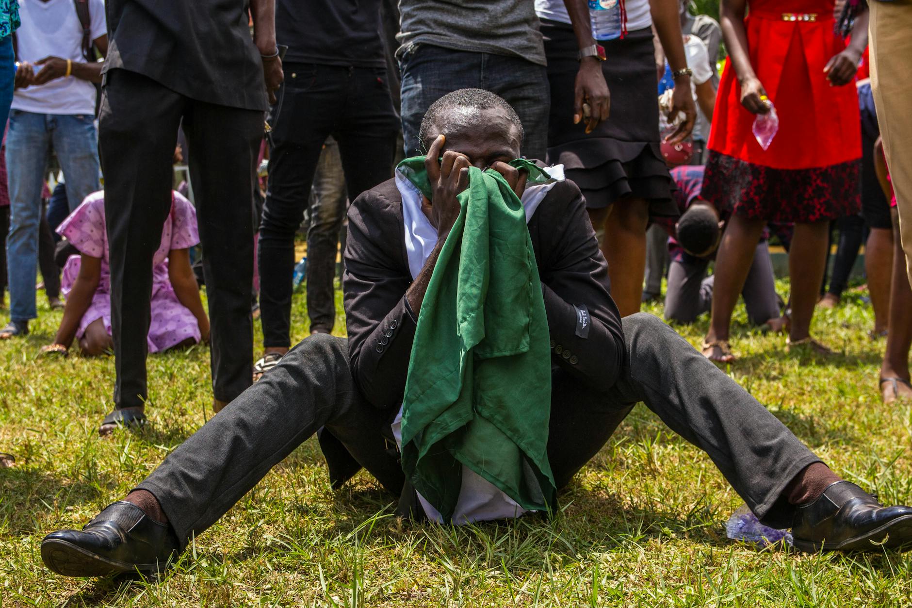 A man sitting on grass in emotional distress, obscured face, surrounded by a crowd outdoors. - spring social anxiety