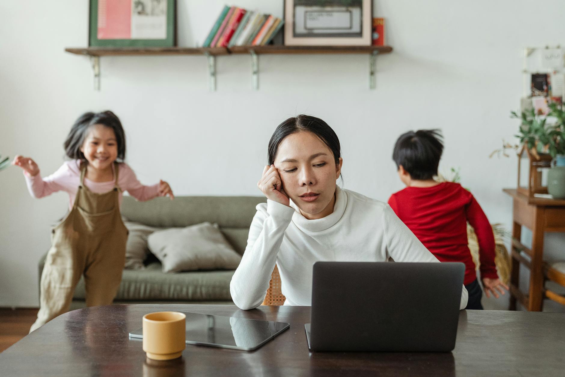 A tired mother working on a laptop while her children play around indoors, highlighting remote work challenges. - spring stress families