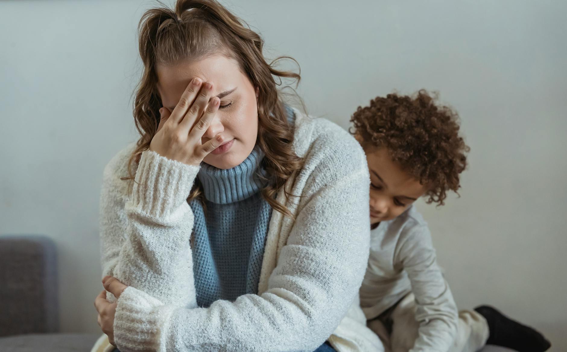 Frustrated mother with hand on forehead and closed eyes sitting near African American son near wall in room at home - spring stress families