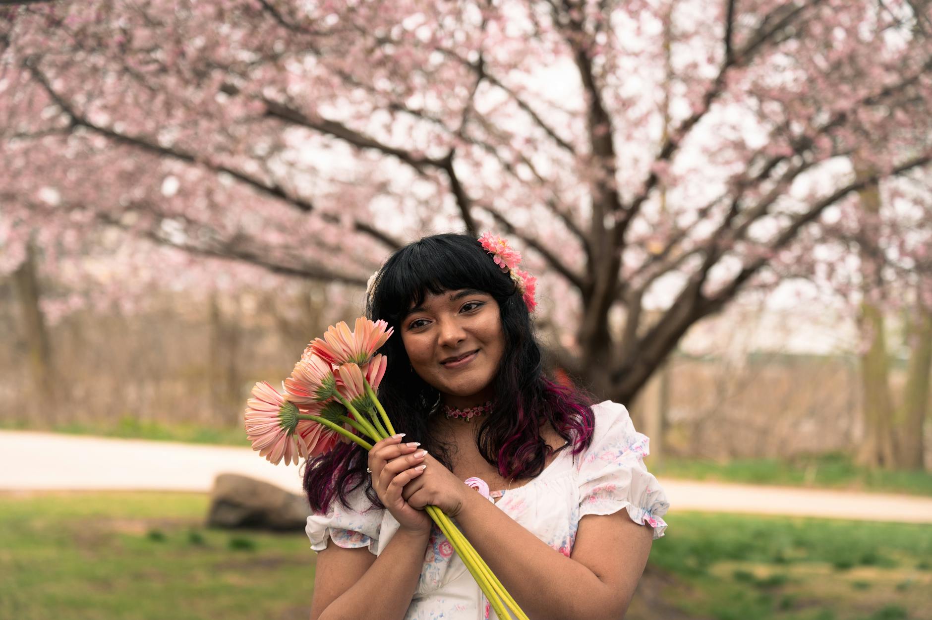 A joyful woman holding pink flowers stands in front of a vibrant cherry blossom tree, capturing a serene spring moment. - springtime sadness
