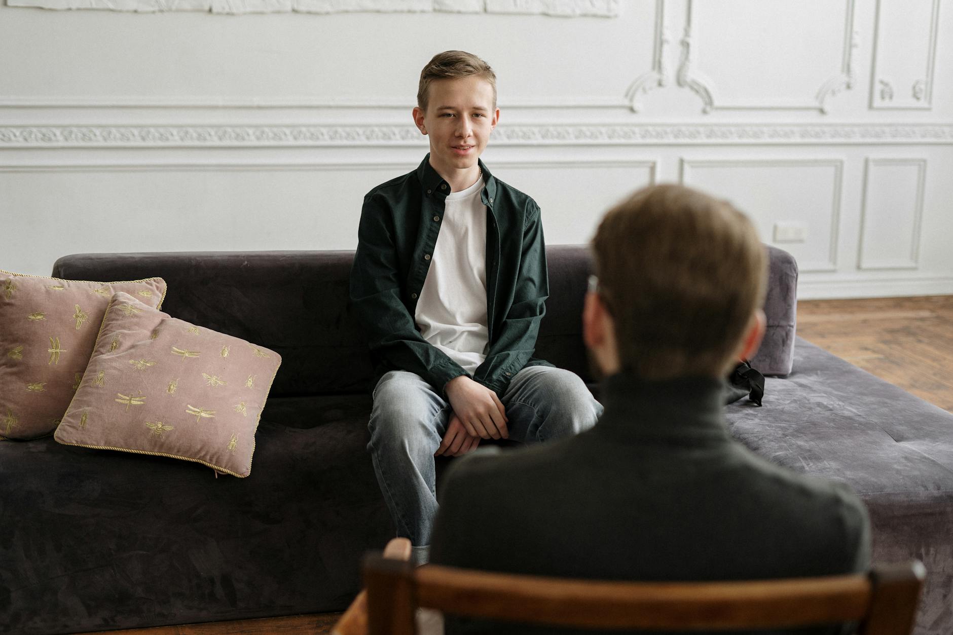 A teenage boy in a counseling session with a therapist in a modern loft setting, sitting on a sofa. - teen mental health