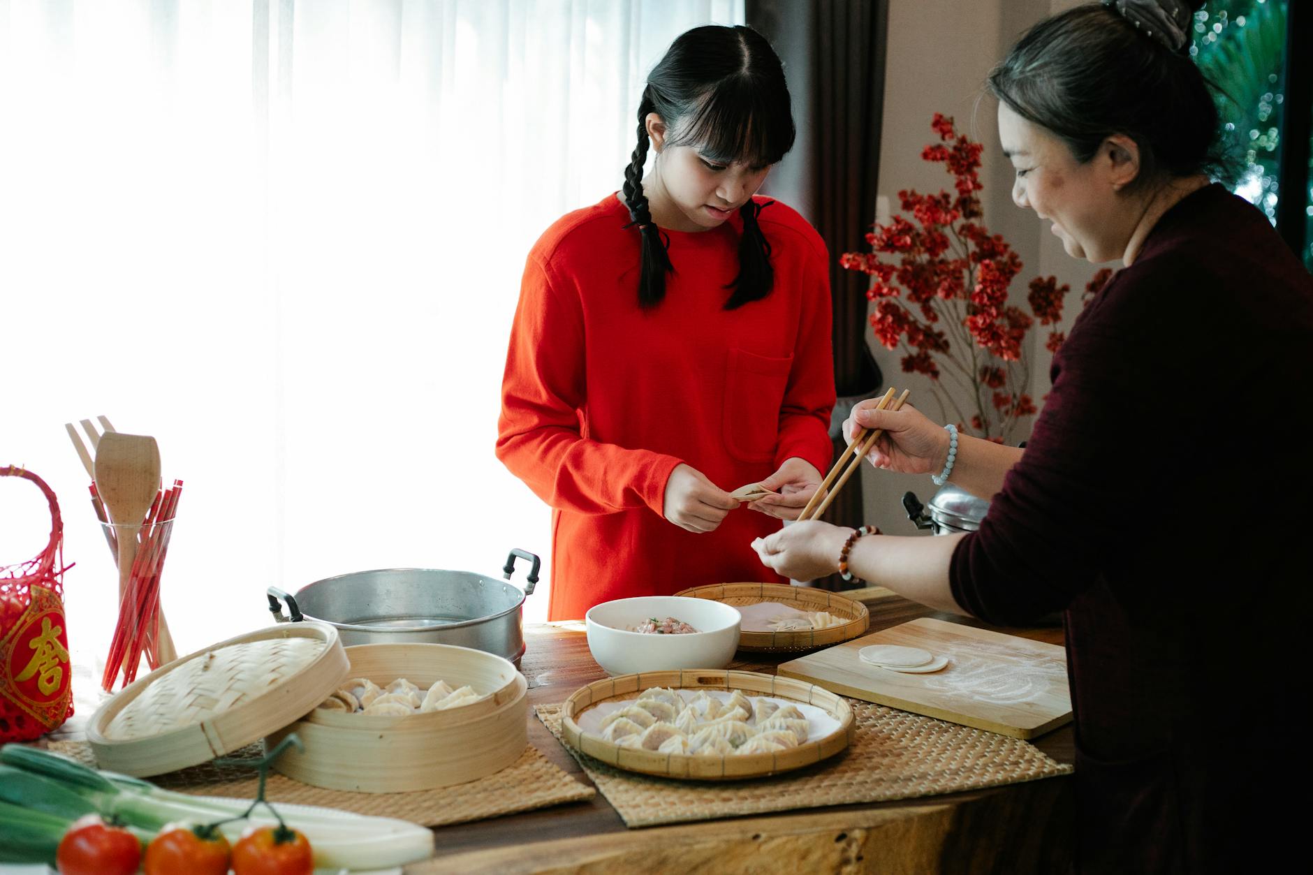 Cheerful ethnic grandma with attentive teen preparing dumplings at table with traditional steamers during New Year holiday at home - teen summer plans