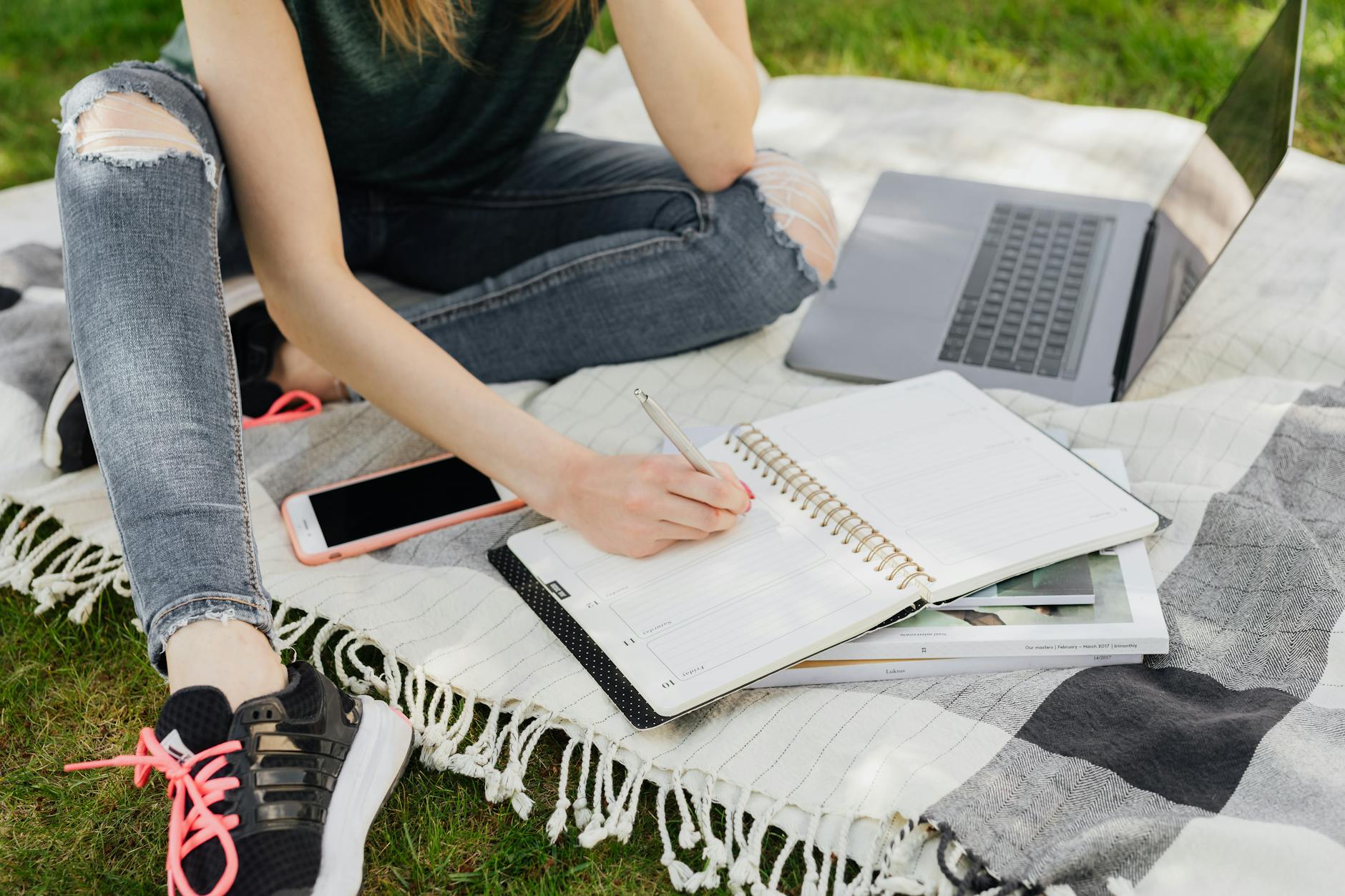 Crop faceless female student in casual clothes taking notes in planner while sitting on blanket with smartphone and laptop on green park grass on sunny day - teen summer plans