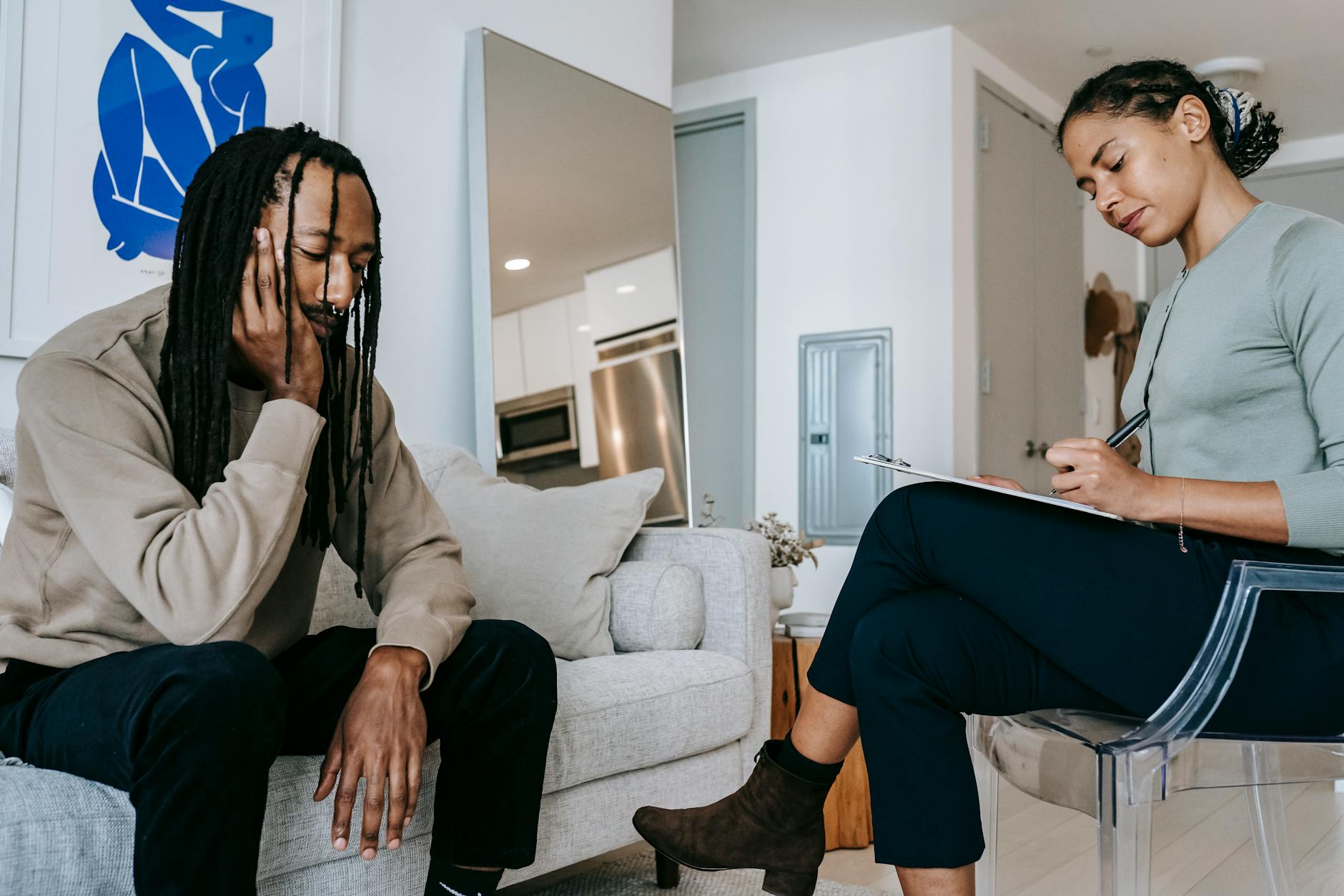 Attentive African American female with clipboard taking notes near sad male partner touching face on couch at home - therapist for seasonal anxiety