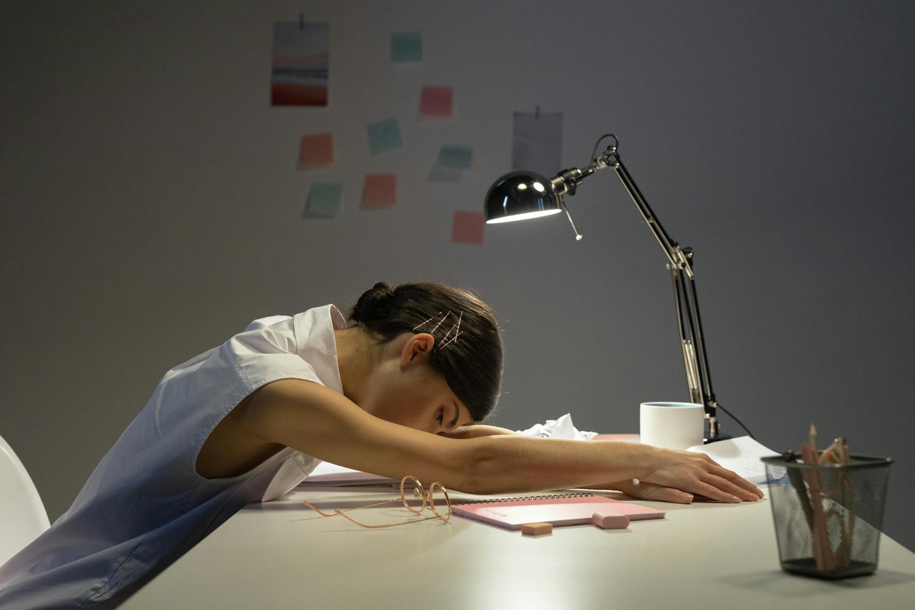 A tired woman rests her head on an office desk under a lamp, symbolizing workplace fatigue. - therapy for burnout