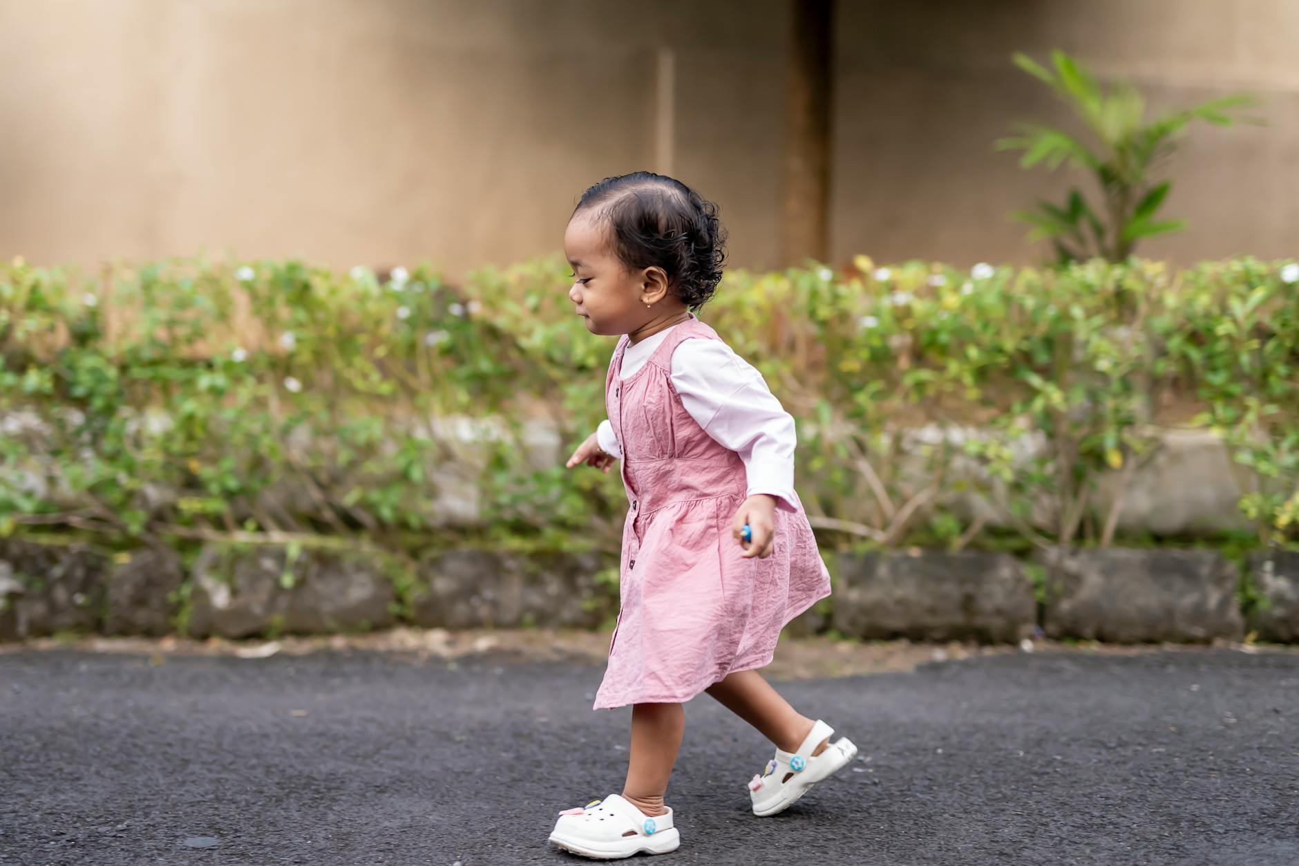 Cute toddler in a pink dress walking on a pathway with green foliage background, showing early steps. - toddler independence winter