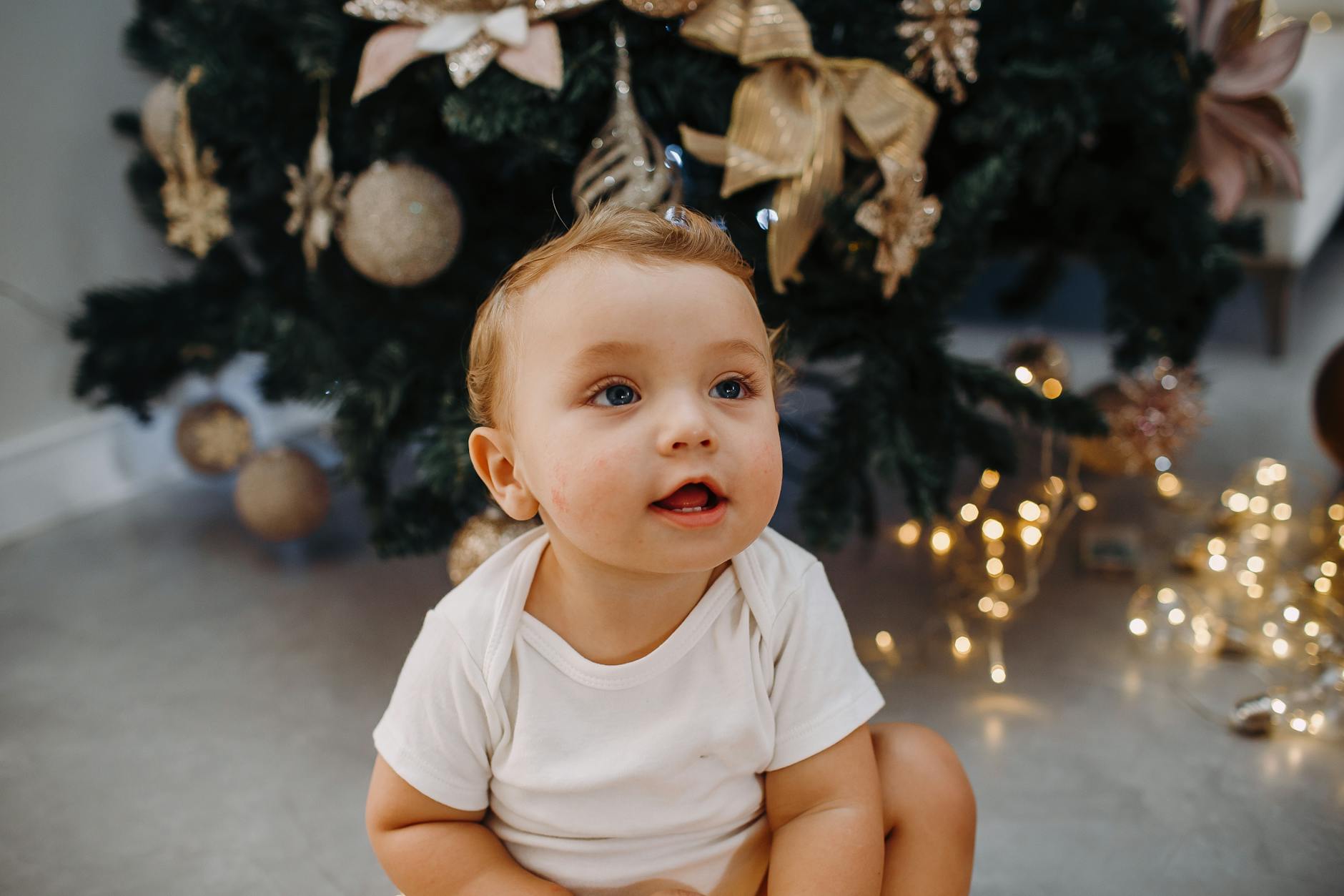 Charming baby in white t-shirt sitting near a beautifully decorated Christmas tree indoors. - toddler independence winter