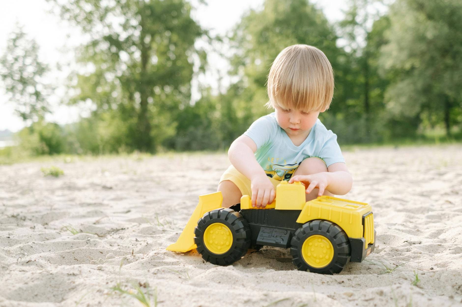 Blonde child enjoys outdoor playtime in a sandy environment with a colorful toy truck. - toddler outdoor independence