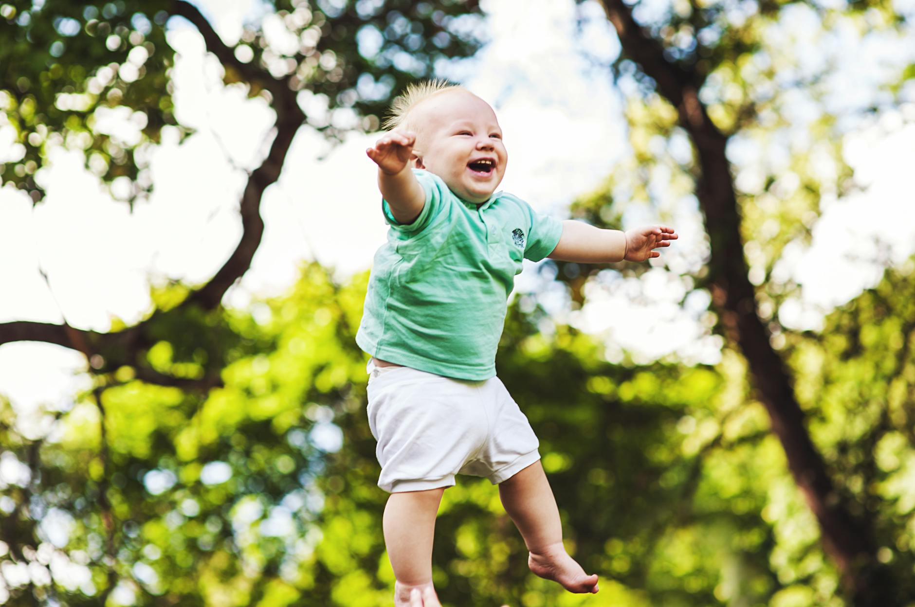 Happy baby wearing a green shirt being tossed in the air on a sunny day in the park. - toddler outdoor play