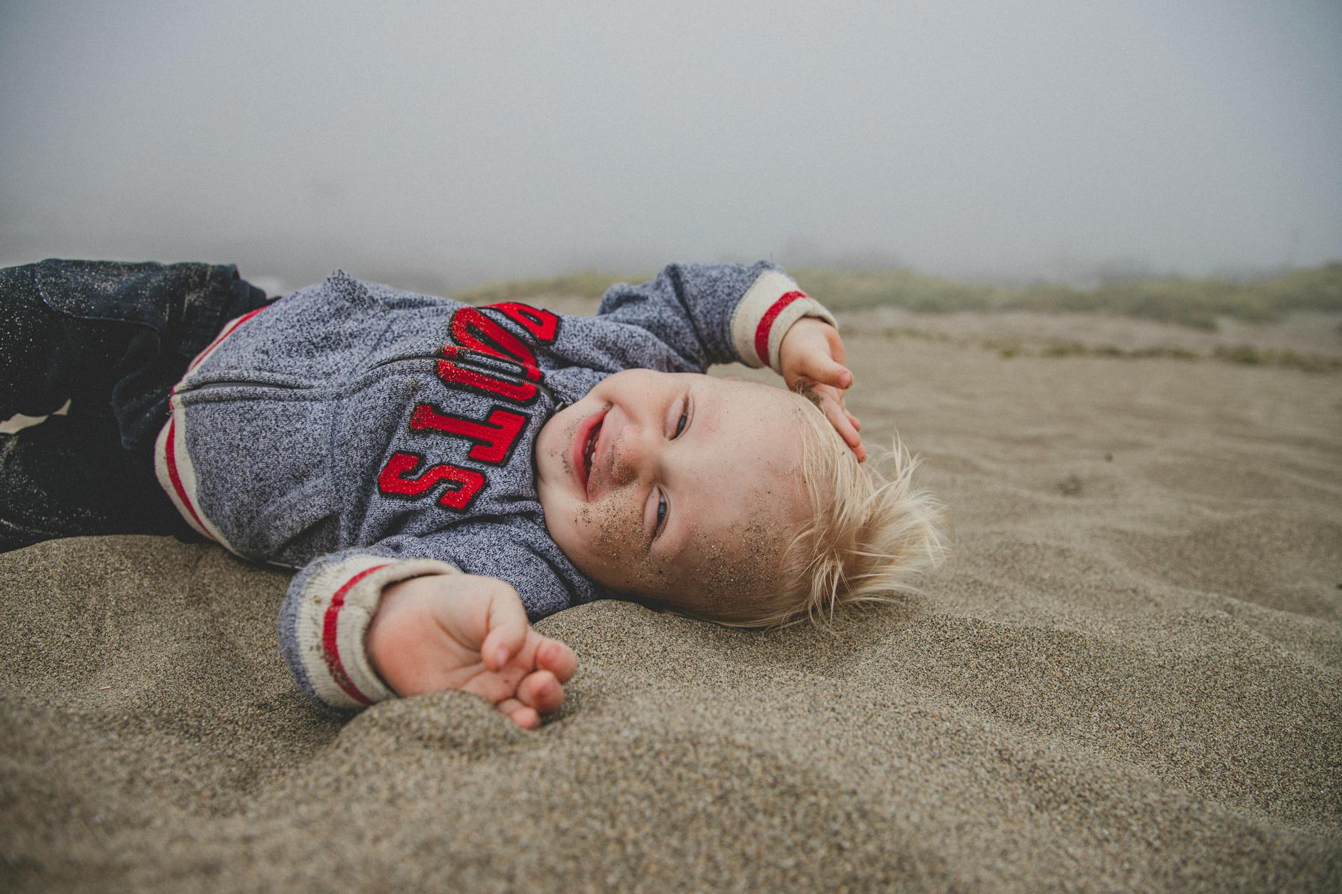 Cute child enjoying a fun day at a foggy beach, lying on the sand and smiling. - toddler outdoor play