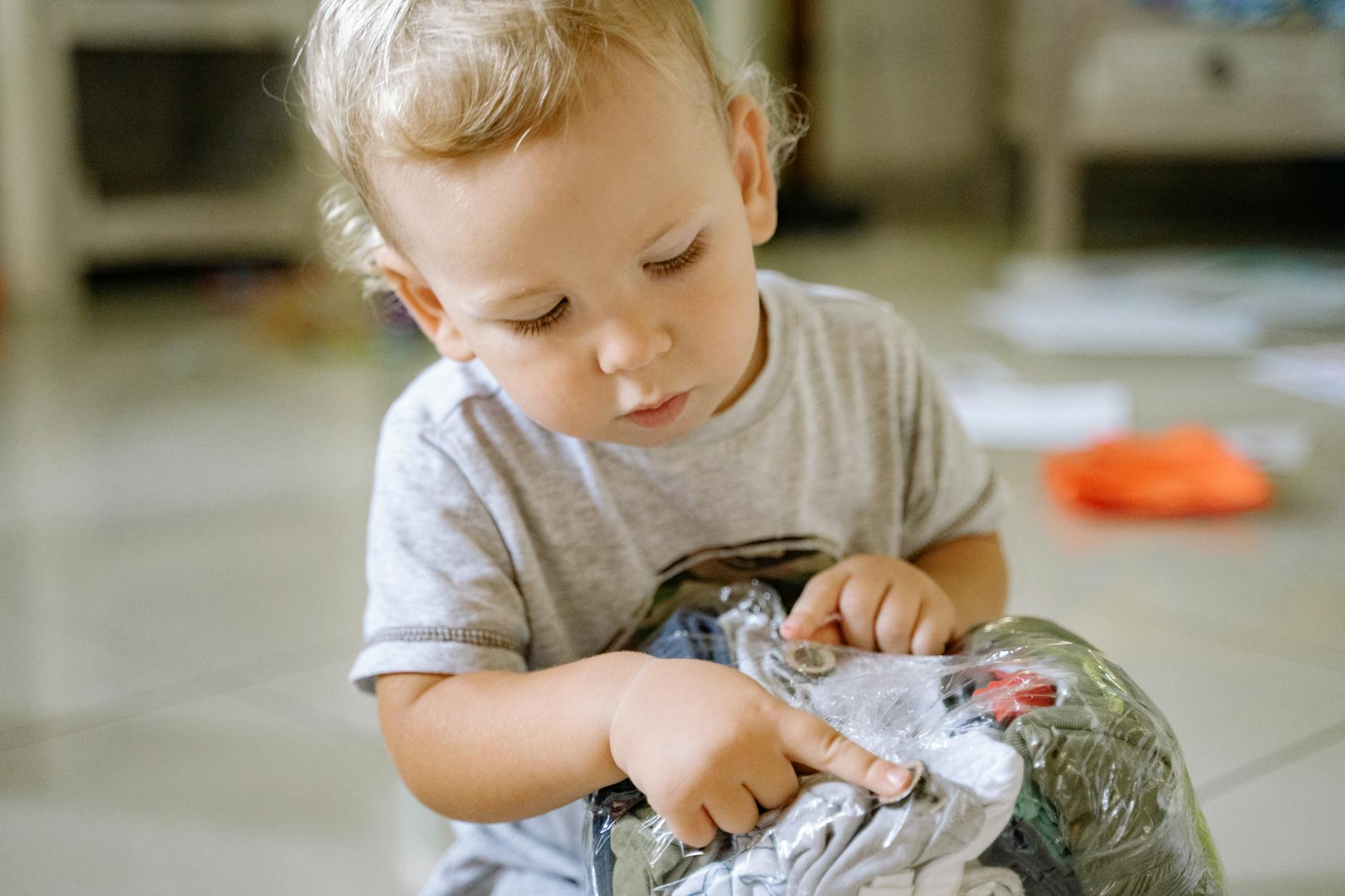 A toddler playing with clothes wrapped in a plastic bag indoors, showcasing curiosity and focus. - toddler sensory play