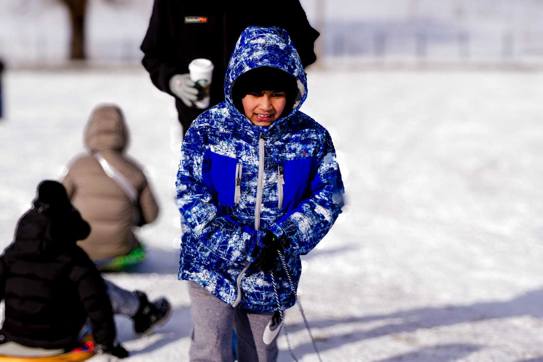 A child in a blue winter jacket playing in the snow with others sledding in the background. - winter anxiety children