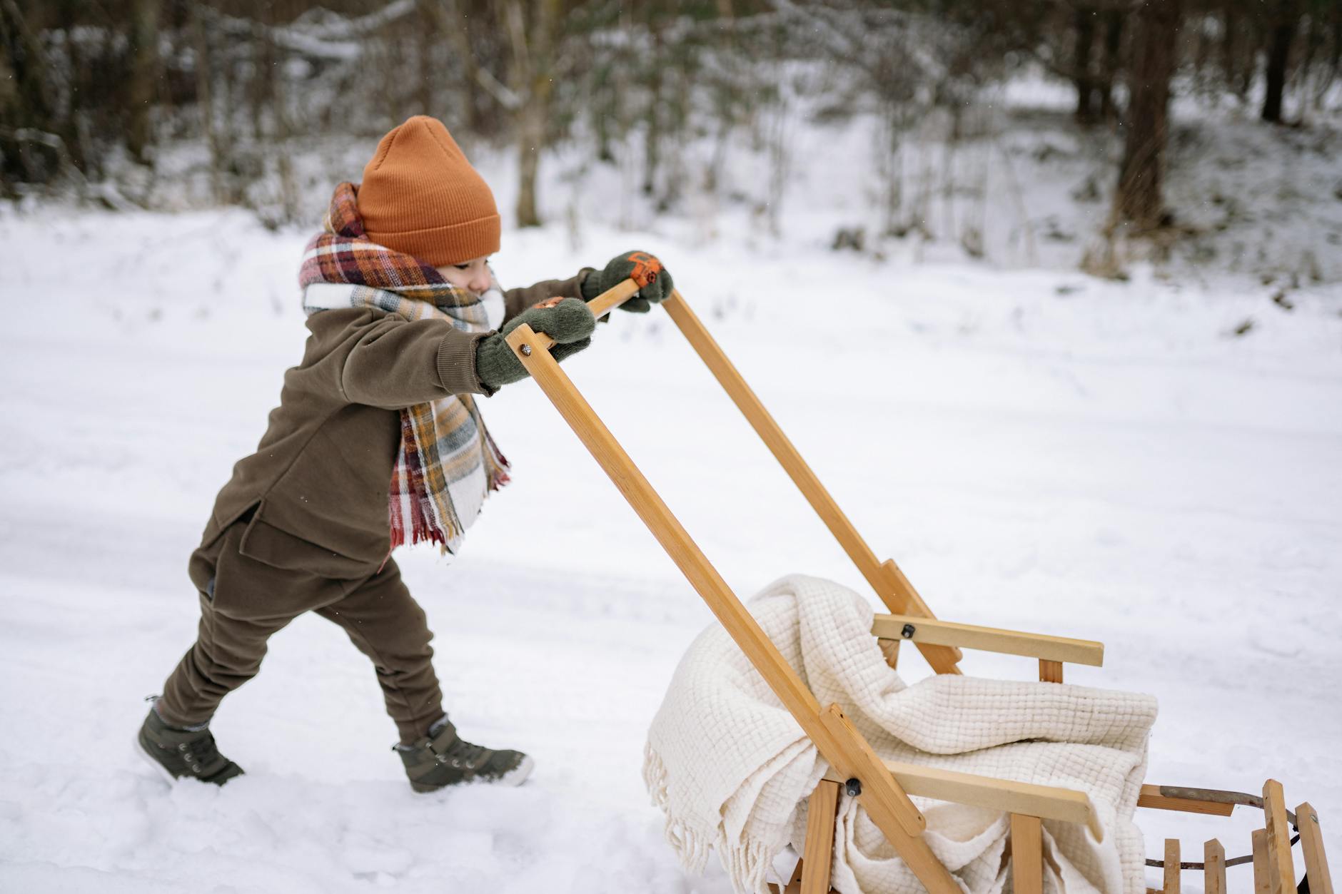 A child dressed warmly pushes a wooden sled through a snowy landscape, enjoying winter outdoor activities. - winter kids activities