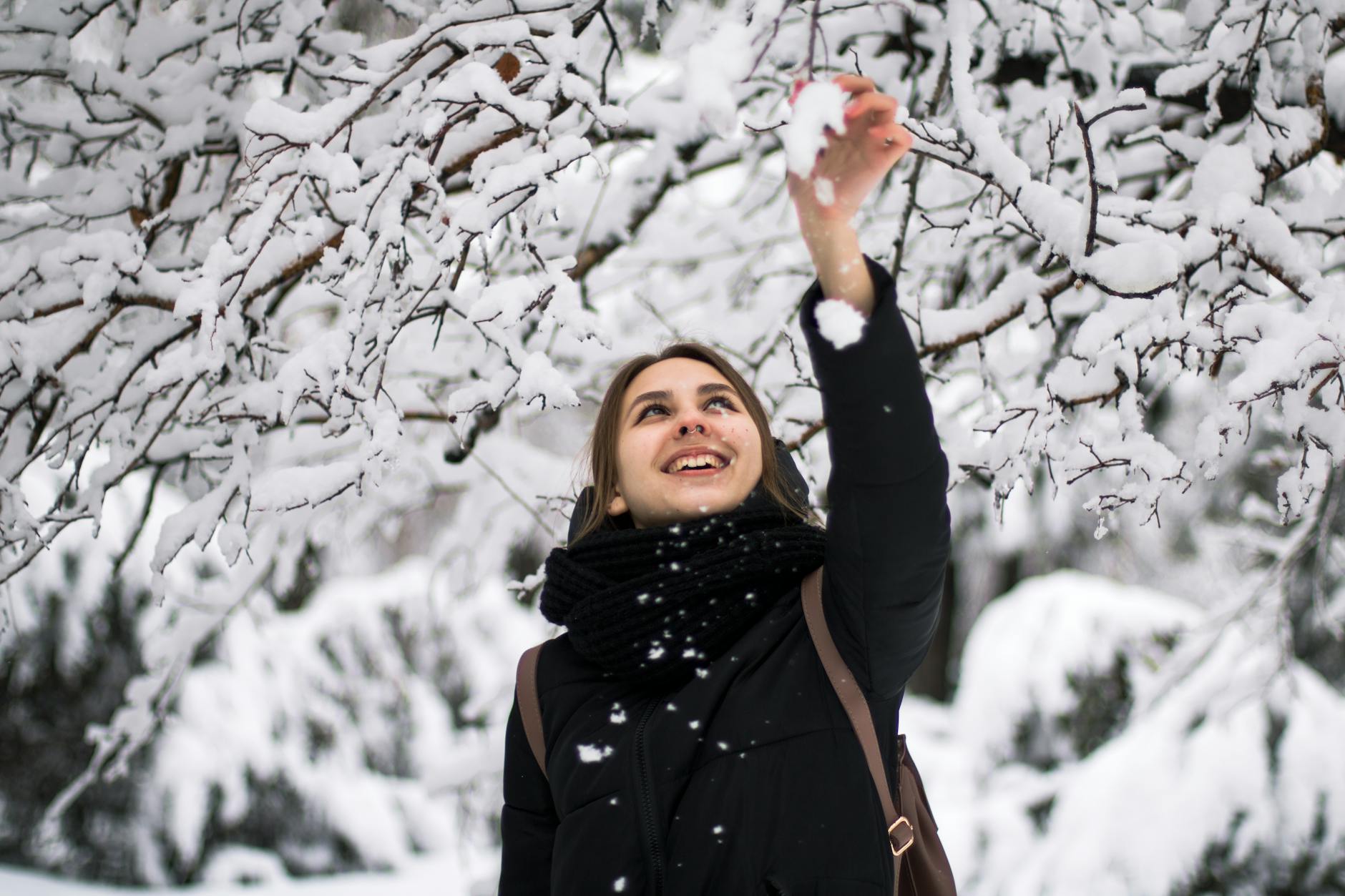A joyful woman reaches for snow on tree branches in a winter wonderland, smiling warmly. - winter sadness therapy