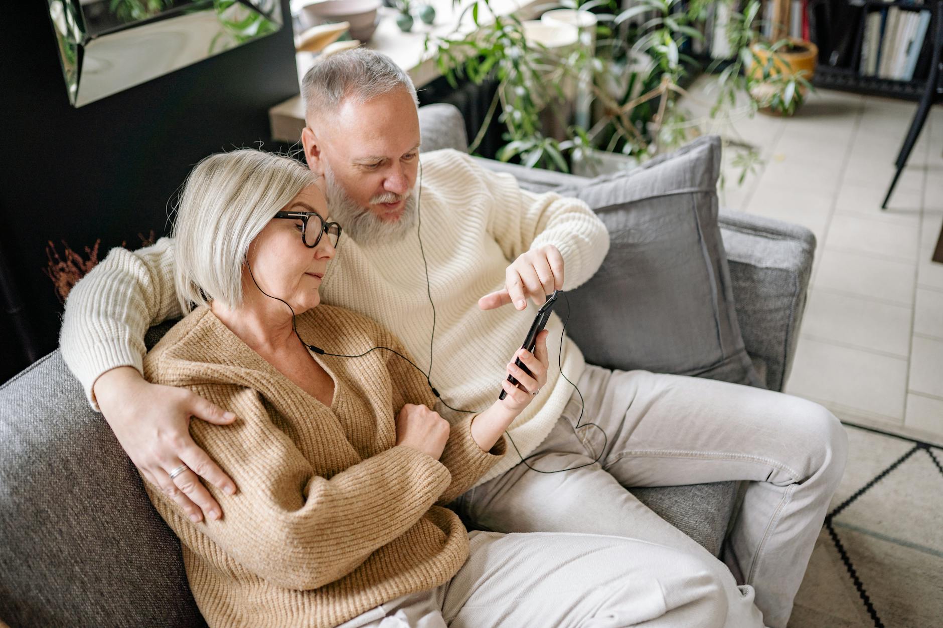 An elderly couple enjoying music on a smartphone while relaxing on a sofa indoors. - active listening couples