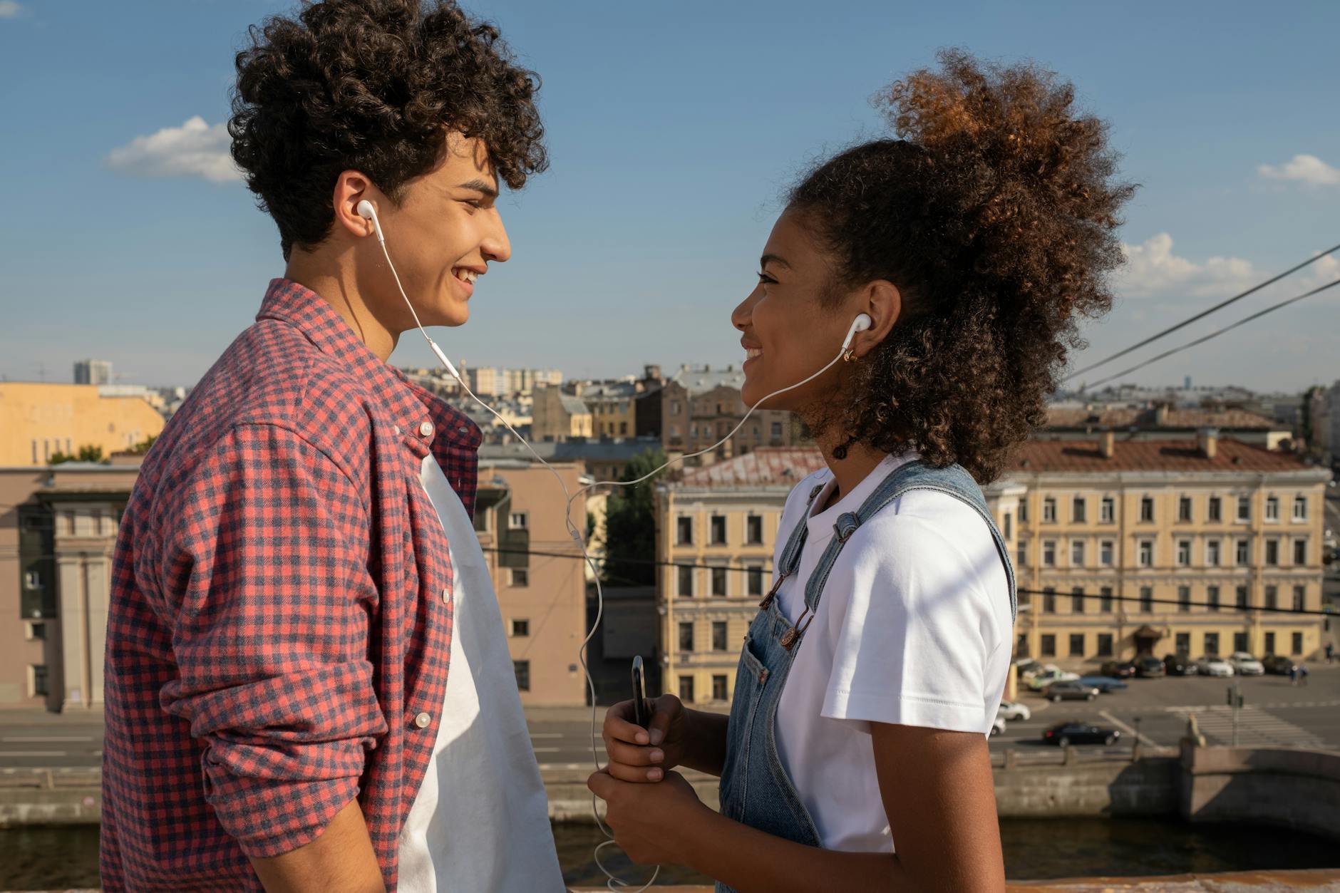 Two teenagers enjoy music together with shared earphones on a sunny rooftop. - active listening couples