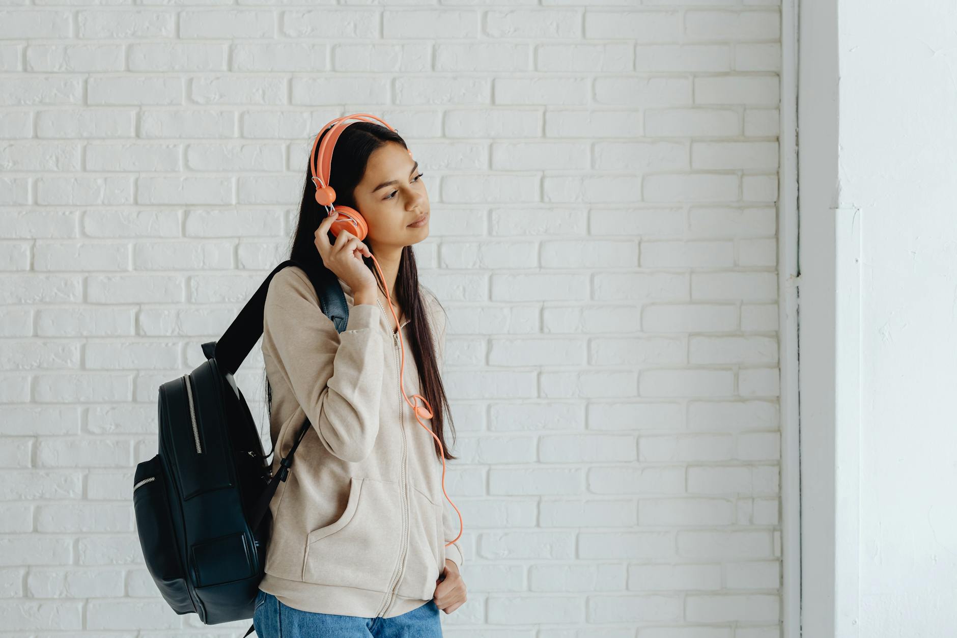 Teen girl with headphones standing against a white brick wall indoors, lost in music. - active listening teen