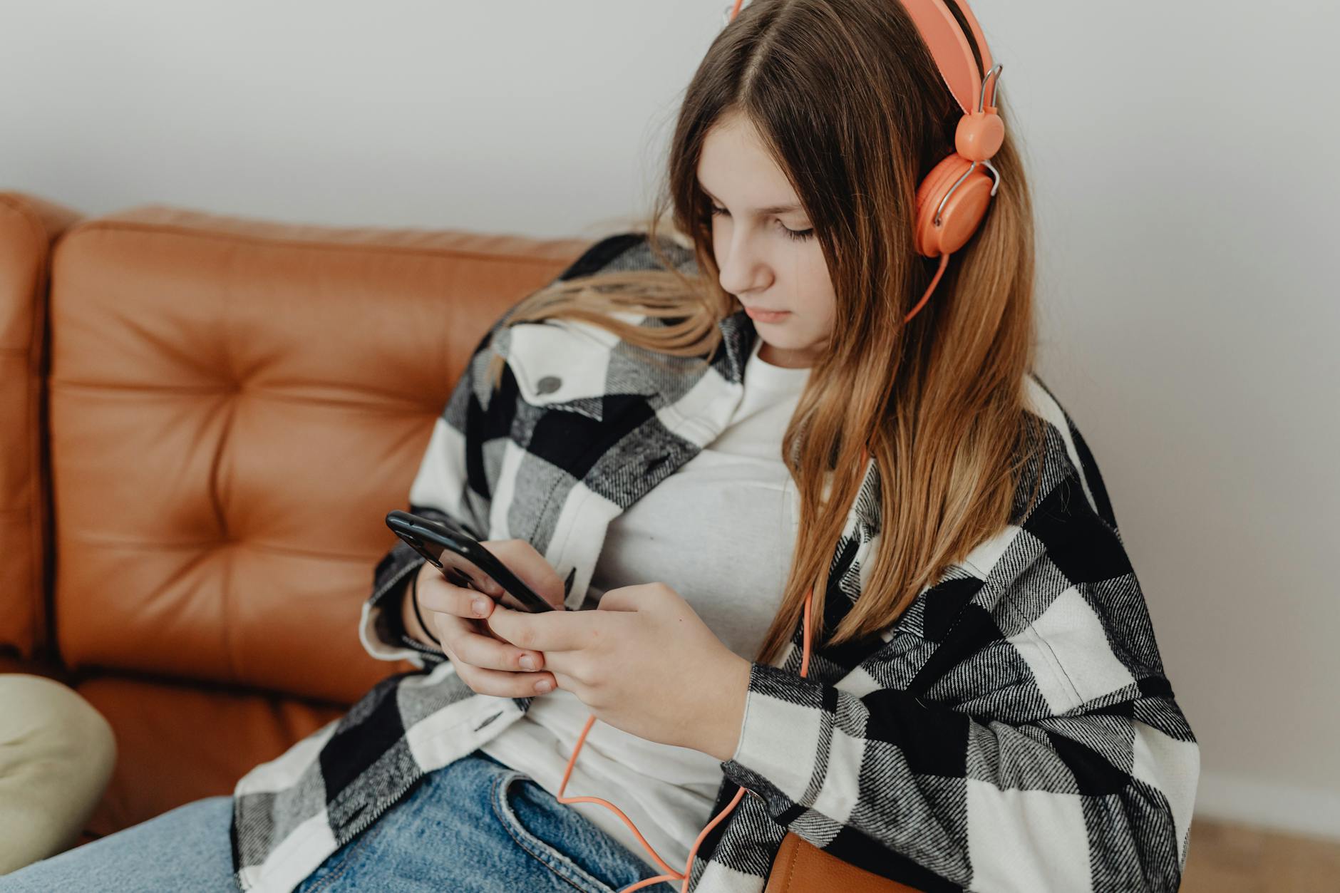 Teen girl sitting on a couch, listening to music with orange headphones, and using a smartphone. - active listening teens