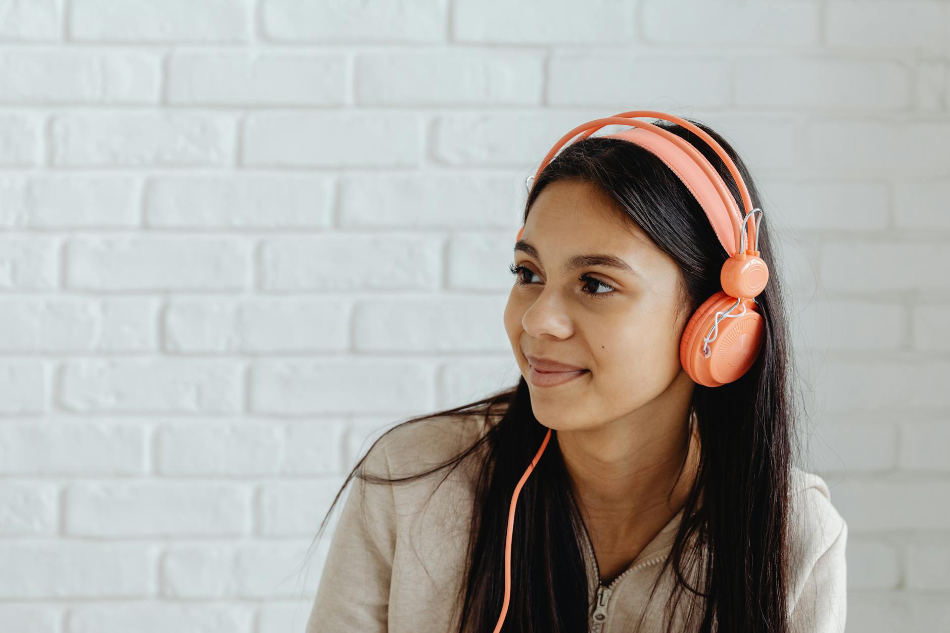 Smiling young woman wearing vibrant headphones, enjoying music indoors against a white brick wall. - active listening teens