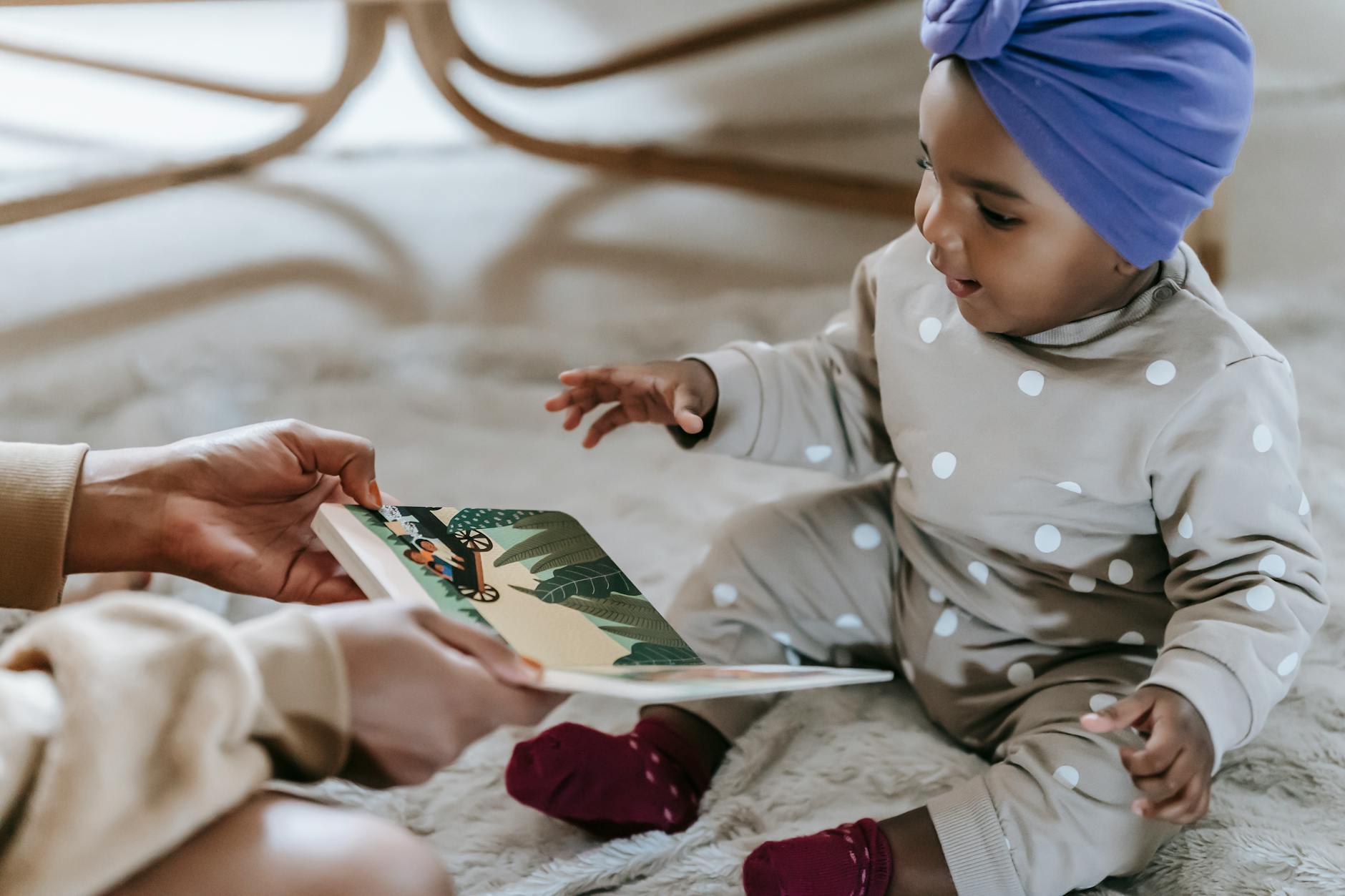 Side view of unrecognizable black mother showing colorful book to exited African American toddler while sitting on floor in light room on blurred background - active listening toddlers