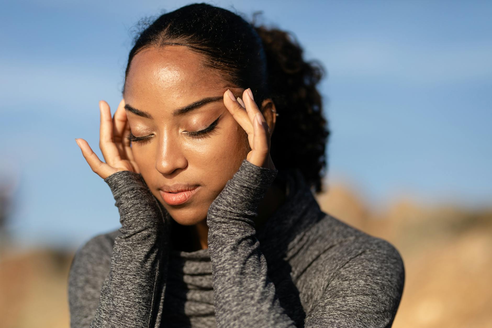 Close-up of a woman in a gray sweater holding her temples, experiencing a headache outdoors in the sunlight. - allergy stress relief