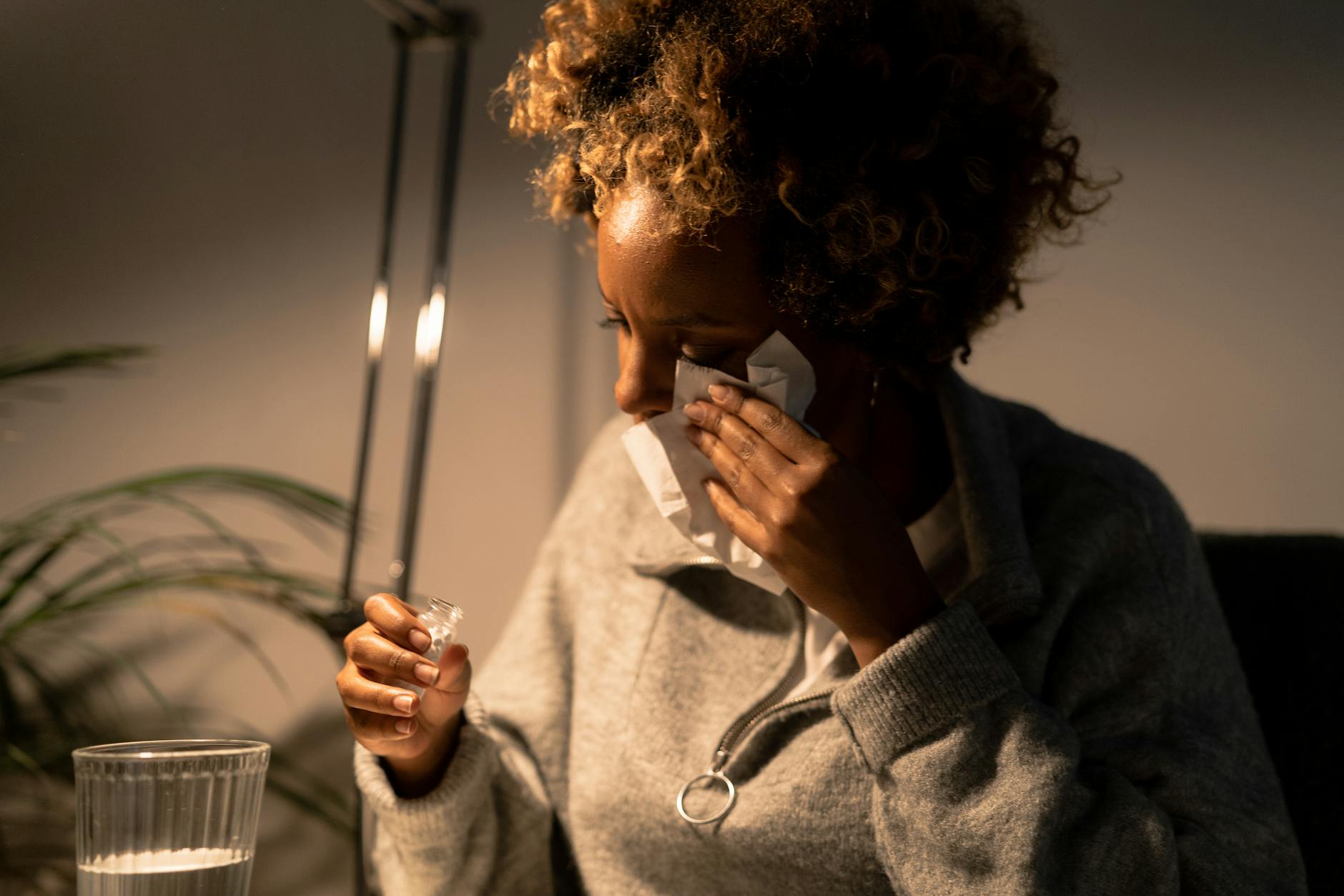 A woman with afro hair wipes tears with a tissue, holding meds and a glass indoors. - allergy stress relief