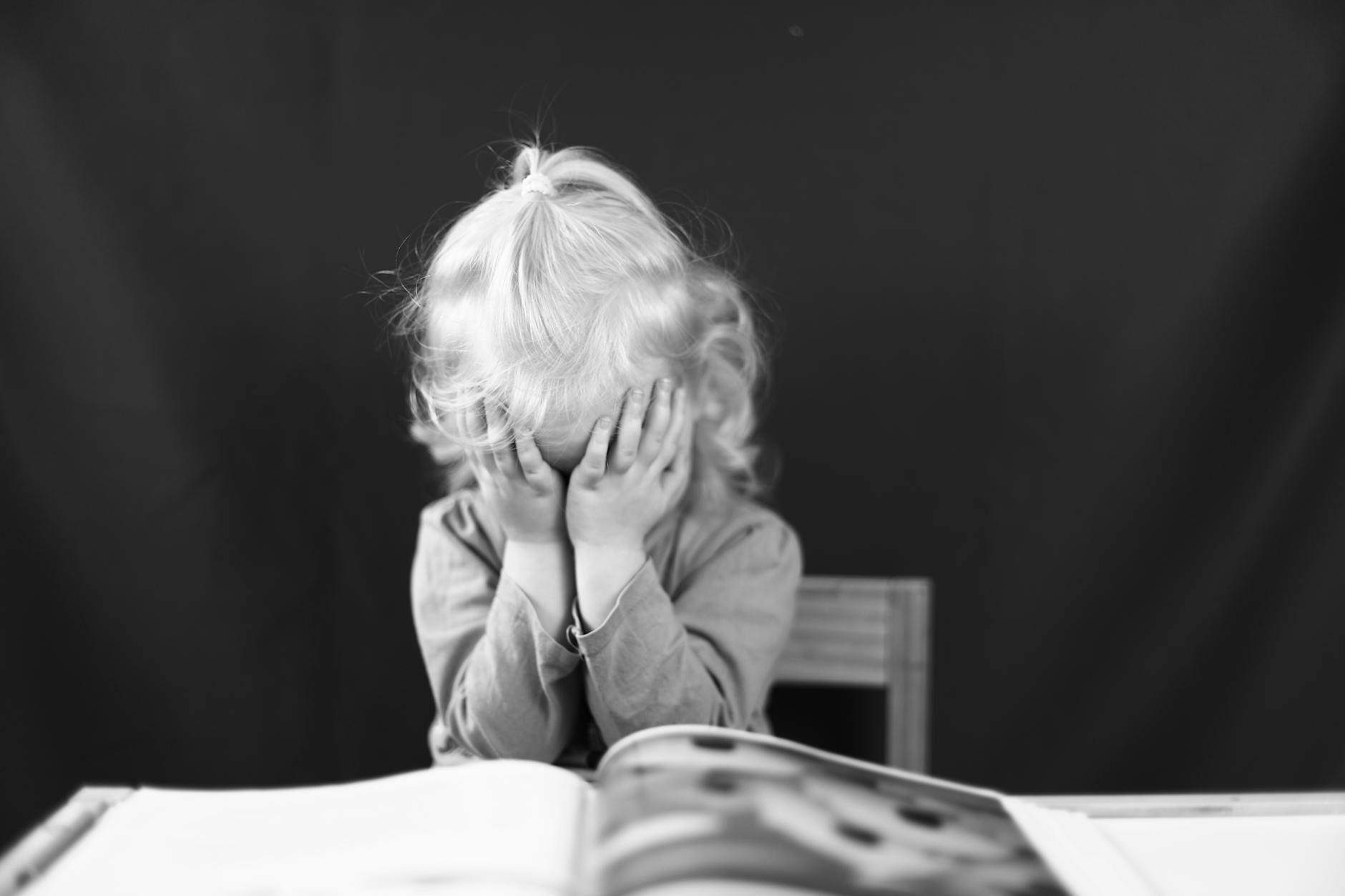 Black and white photo of a young child covering their face with hands, sitting in front of a book. - child emotional changes