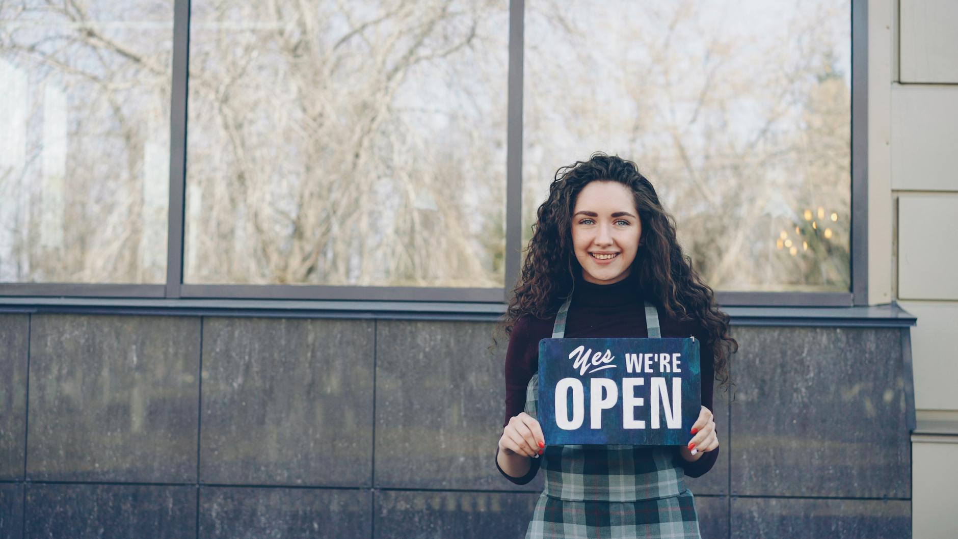 Happy woman holding 'Yes We're Open' sign in front of a store. - communicate needs busy season