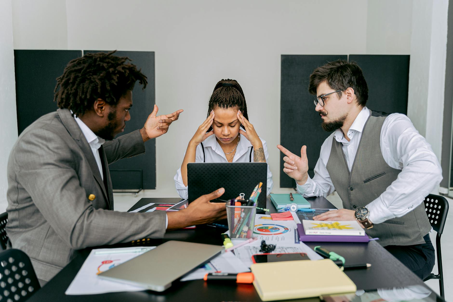 Two men argue while a woman looks frustrated at a laptop in an office environment. - conflict meaning