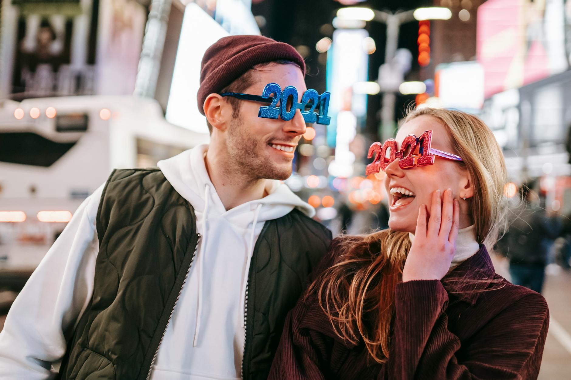 Smiling couple wearing decorated 2021 glasses looking at each other while standing on street with illuminated buildings on blurred background - couple resolutions